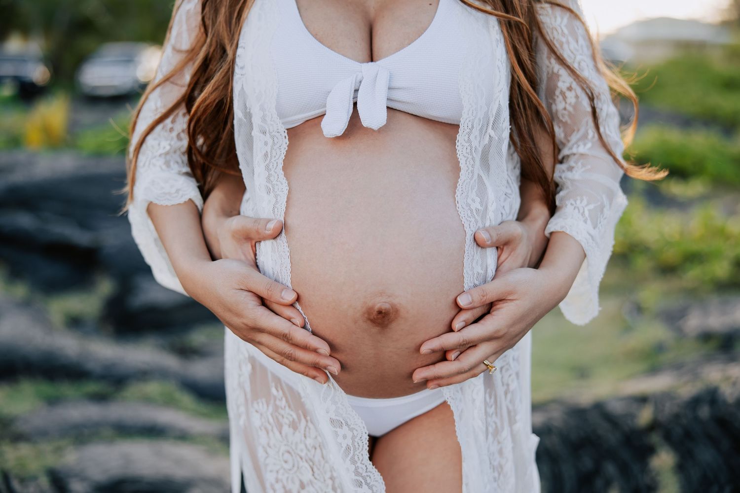 Pregnant woman in white lace robe and bikini cradling her bare baby bump outdoors in natural light.