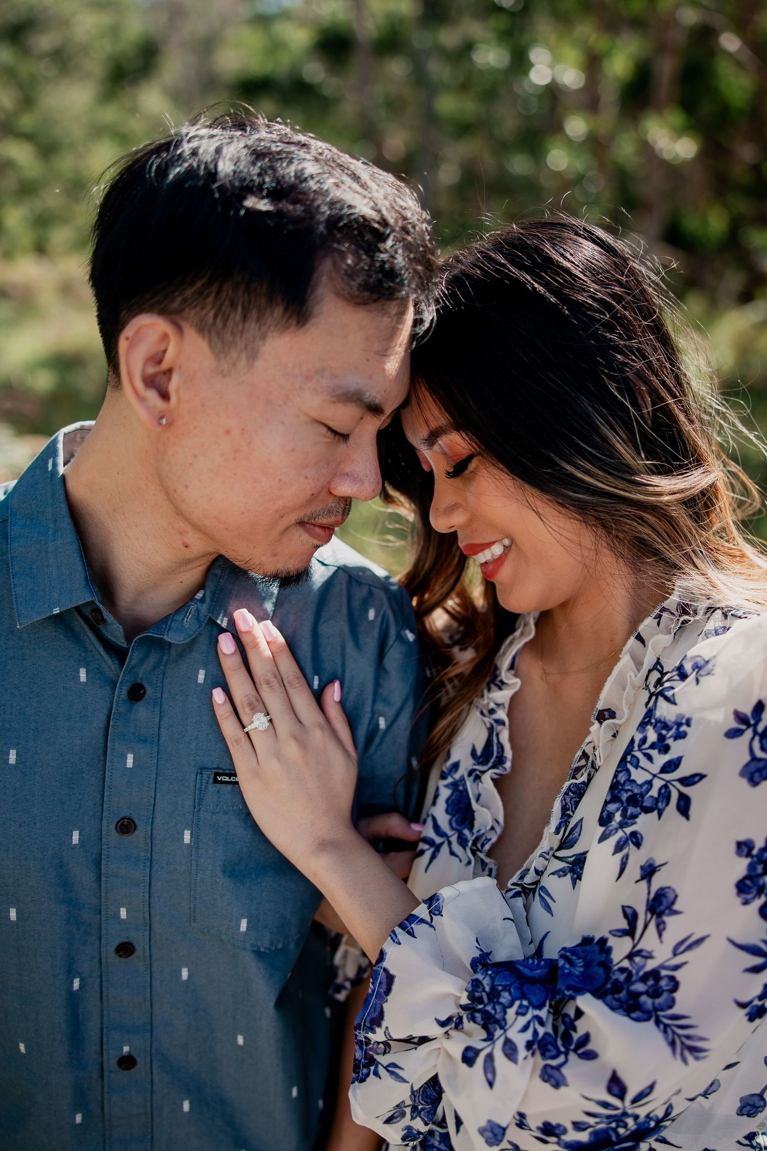 Couple sharing a romantic forehead touch outdoors, woman in floral dress showing engagement ring.