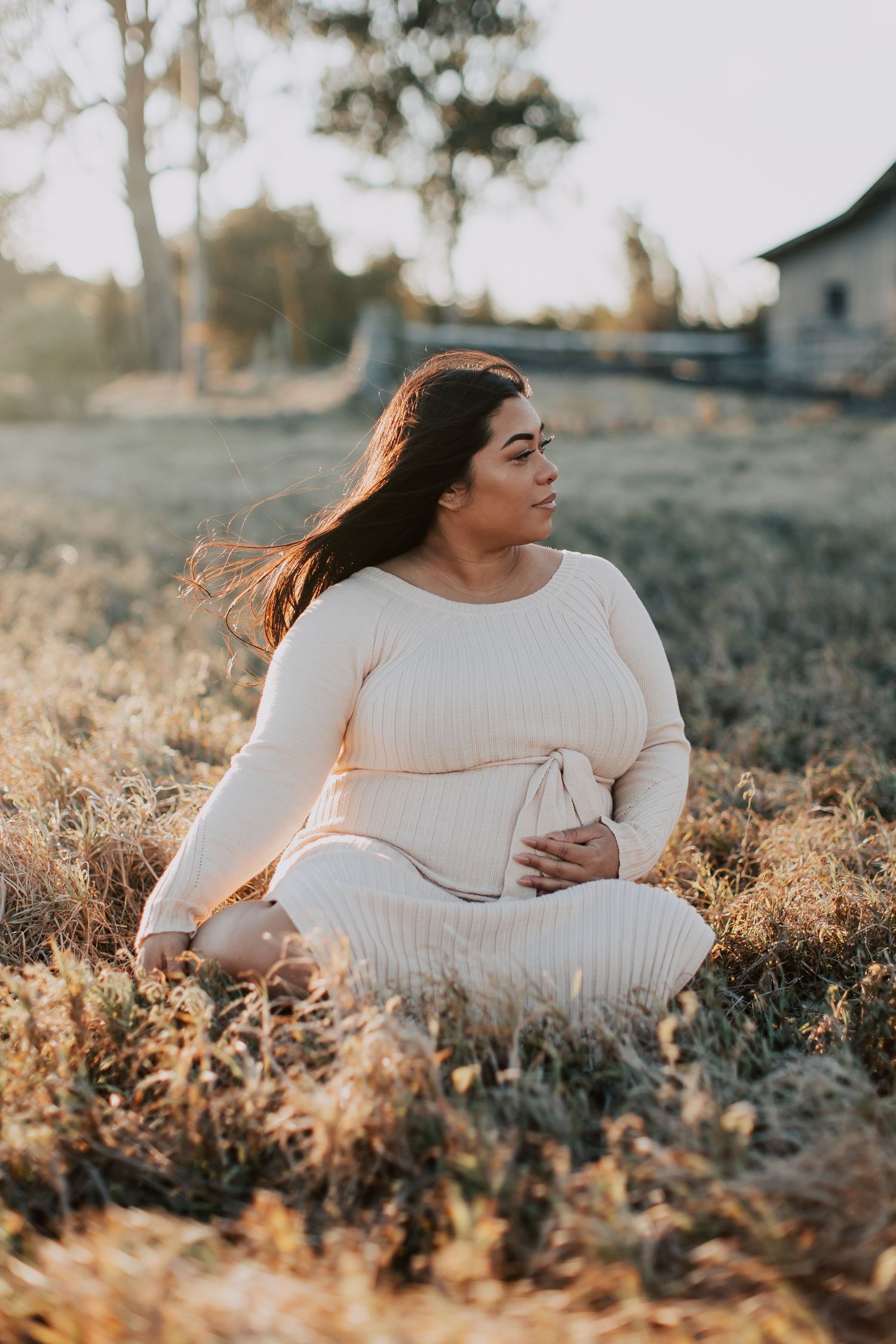 Pregnant woman in cream ribbed dress sitting cross-legged in golden field at sunset, cradling baby bump.