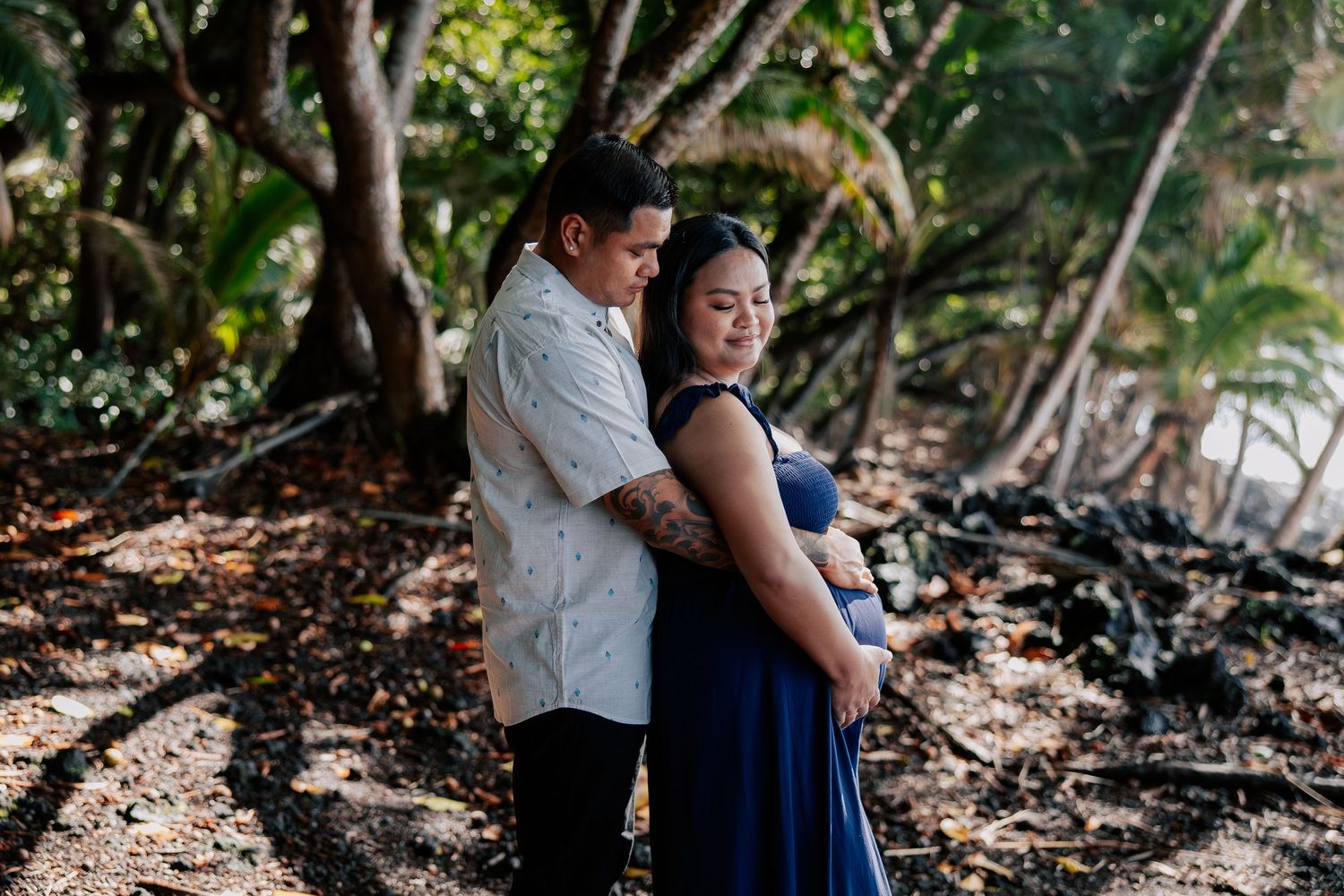 Couple embracing in a tropical forest setting, woman in blue maxi dress, man in white shirt, dappled sunlight.
