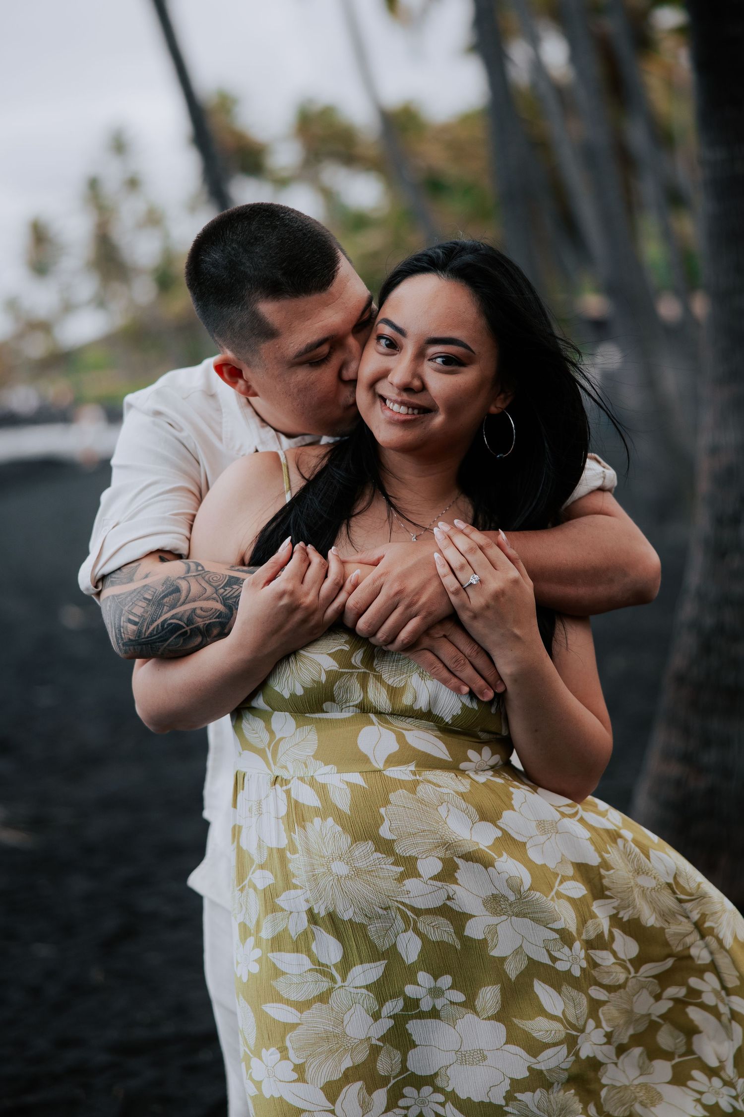 A couple embraces on a black sand beach in Hawaii, the man kissing her cheek as she smiles showing her engagement ring.