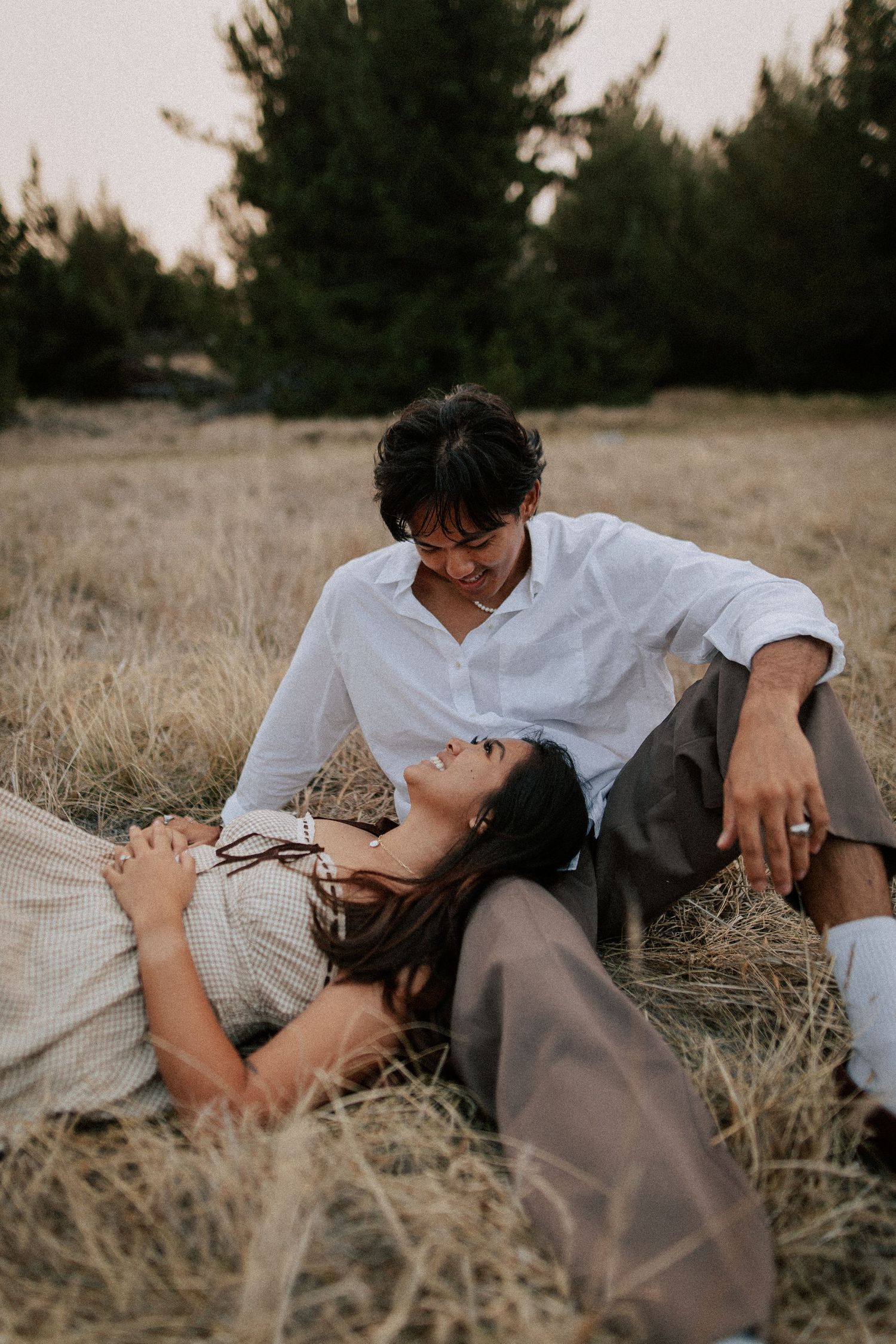 Couple sharing a romantic moment in a golden field, woman lying in grass laughing as man gazes down at her.