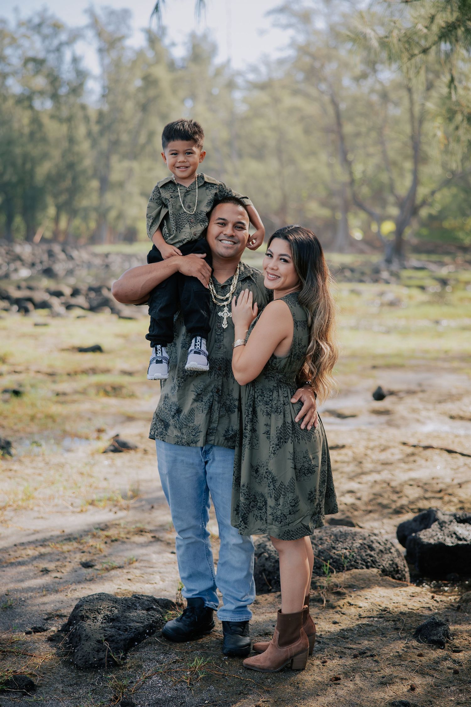 Family of three posing outdoors, parents in matching olive outfits with young child on dad's shoulders, smiling.