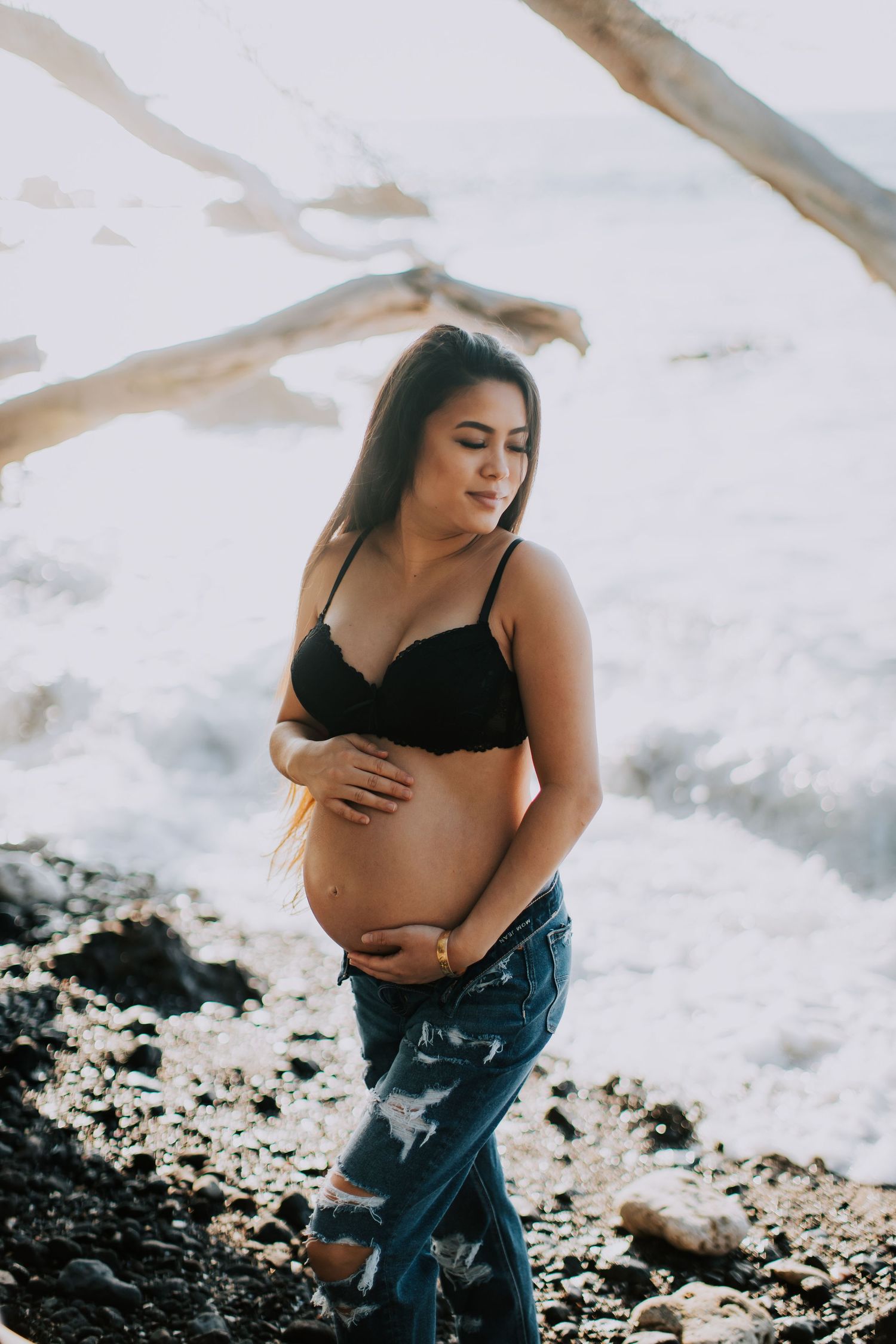 Pregnant woman in black bra and ripped jeans cradling baby bump on rocky beach with ocean waves.