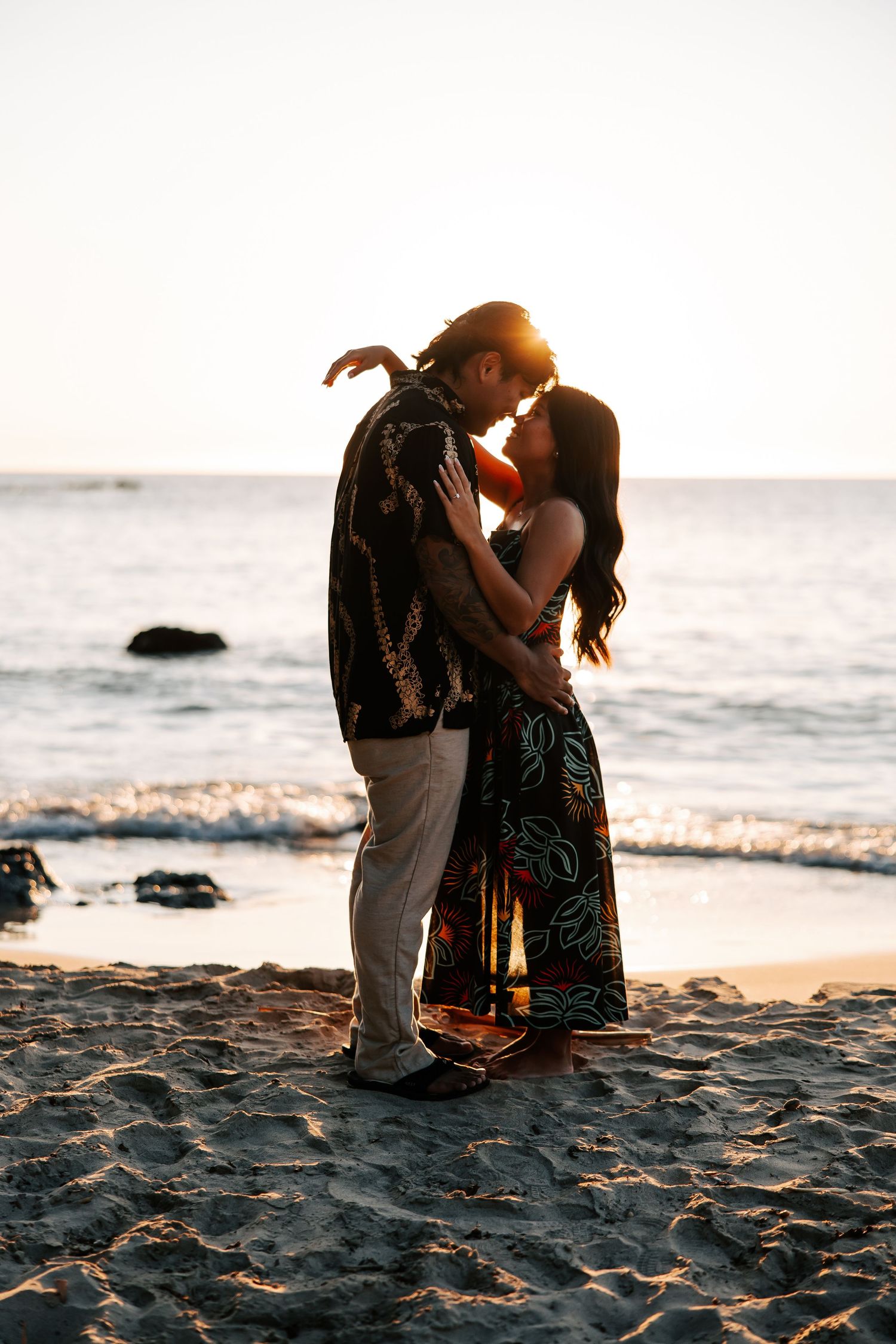 Couple sharing a romantic kiss on a sandy beach at golden sunset with ocean waves in the background.