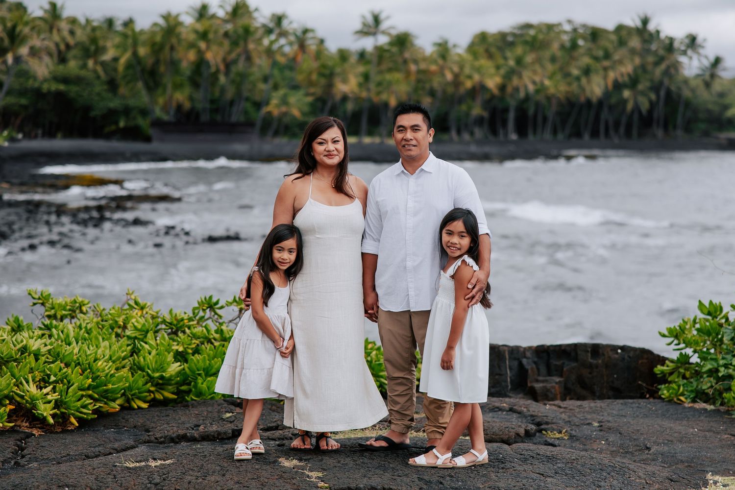 Family of four in white outfits posing on black lava rock by a tropical ocean coastline in Hawaii.