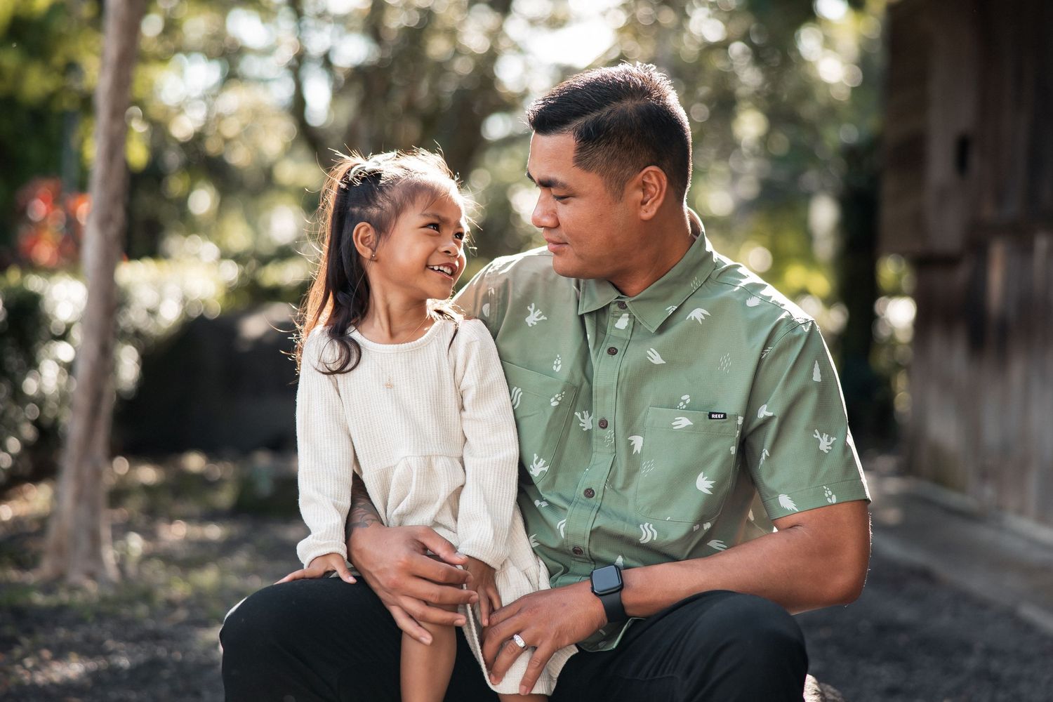 A man and young girl share a joyful moment outdoors, smiling at each other in a sunlit park setting.