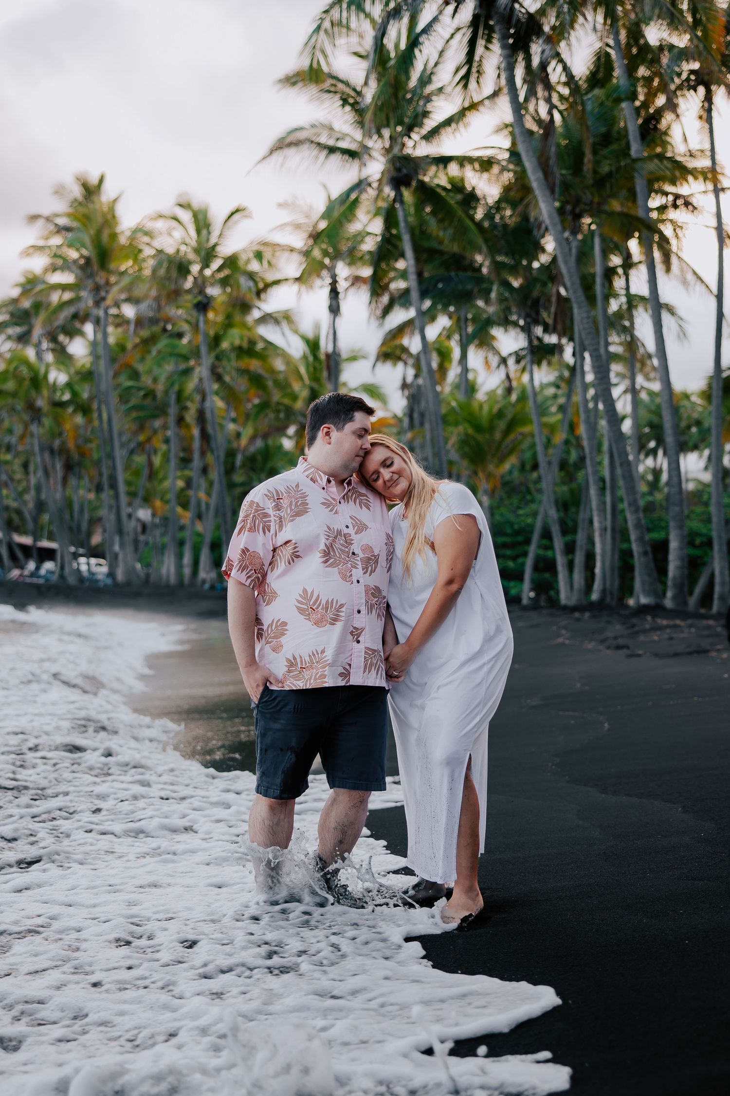 Couple standing on black sand beach with palm trees, waves washing over their feet in a romantic tropical setting.