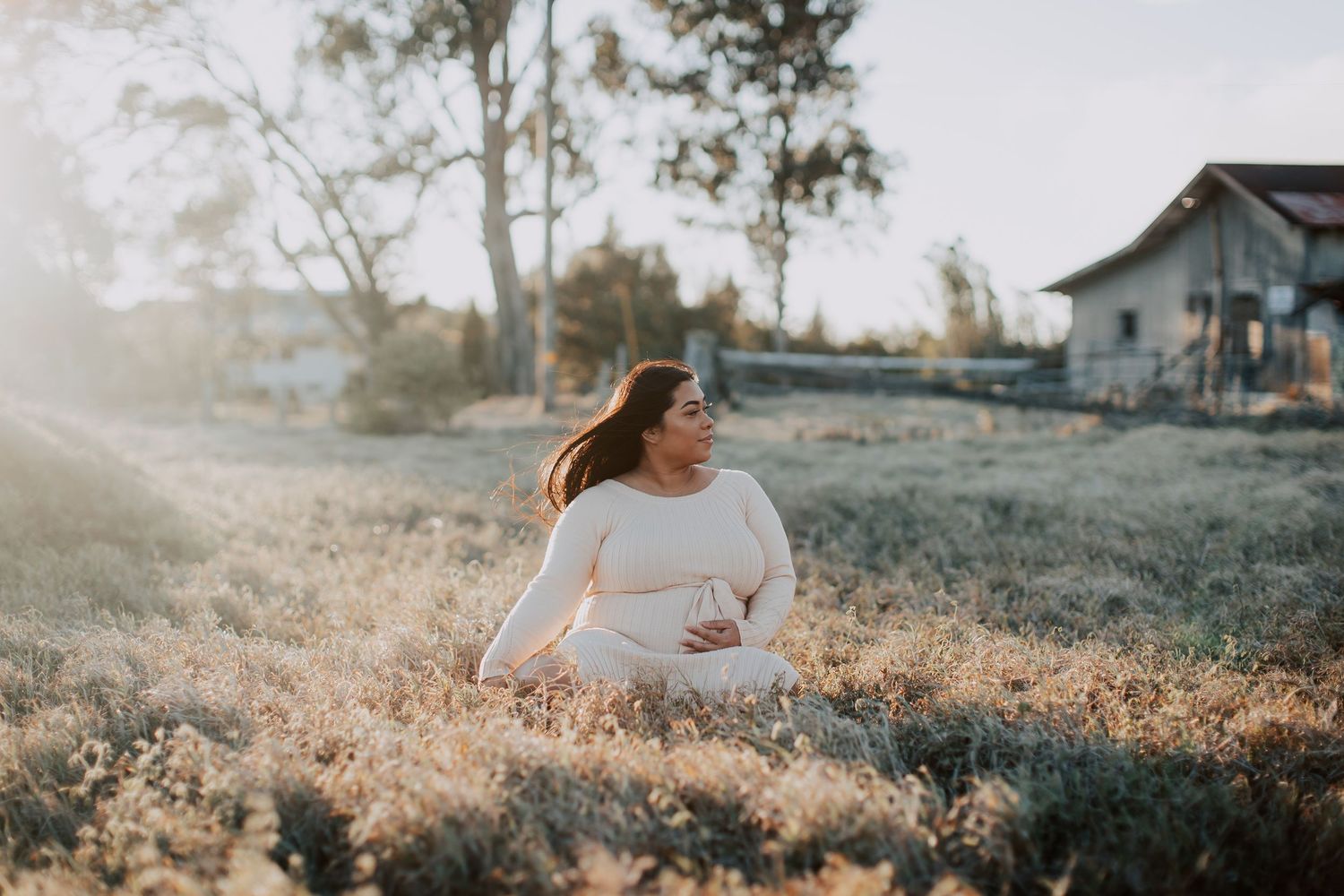 Pregnant woman in white outfit sitting in golden field at sunset near rustic barn.