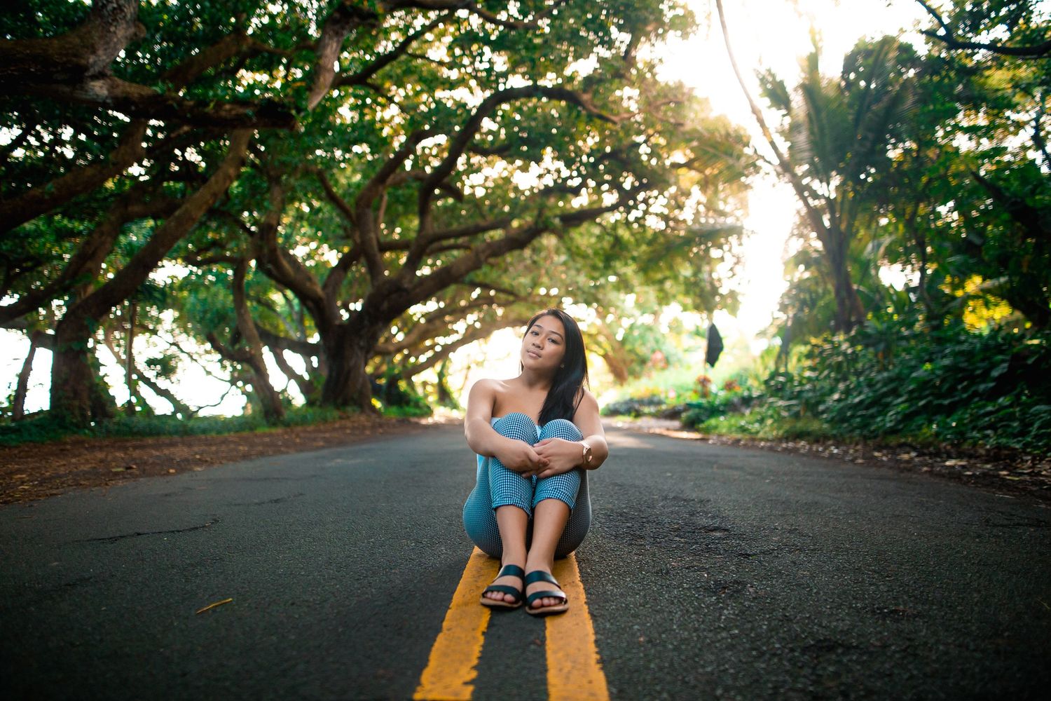 A young woman sits cross-legged on a tree-lined road, smiling upward in golden sunlight.