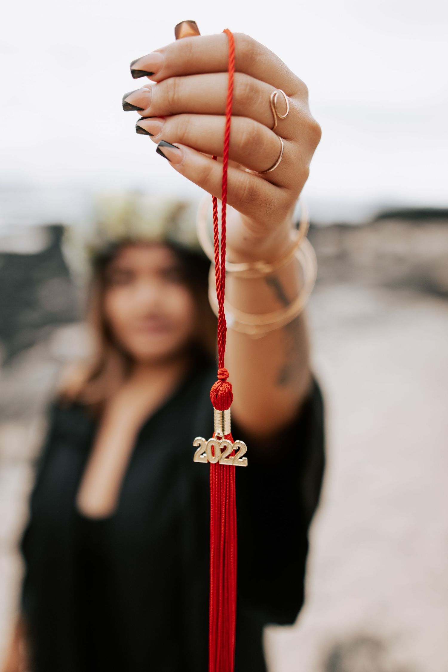 Woman holding a red 2022 graduation tassel, wearing dark nail polish and gold rings, with blurred background.