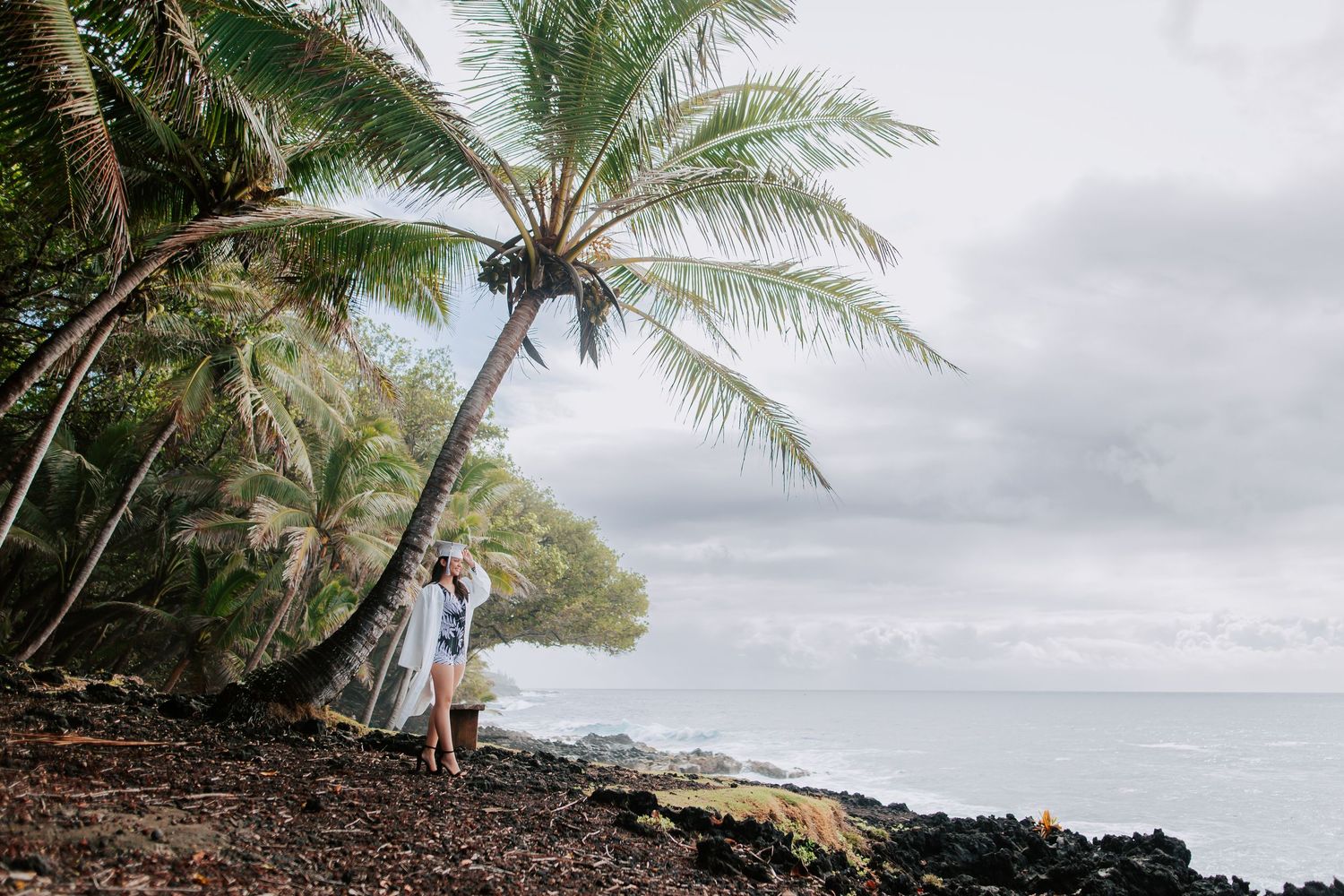 Couple embracing under a leaning palm tree on a rocky Hawaiian coastline with ocean views.
