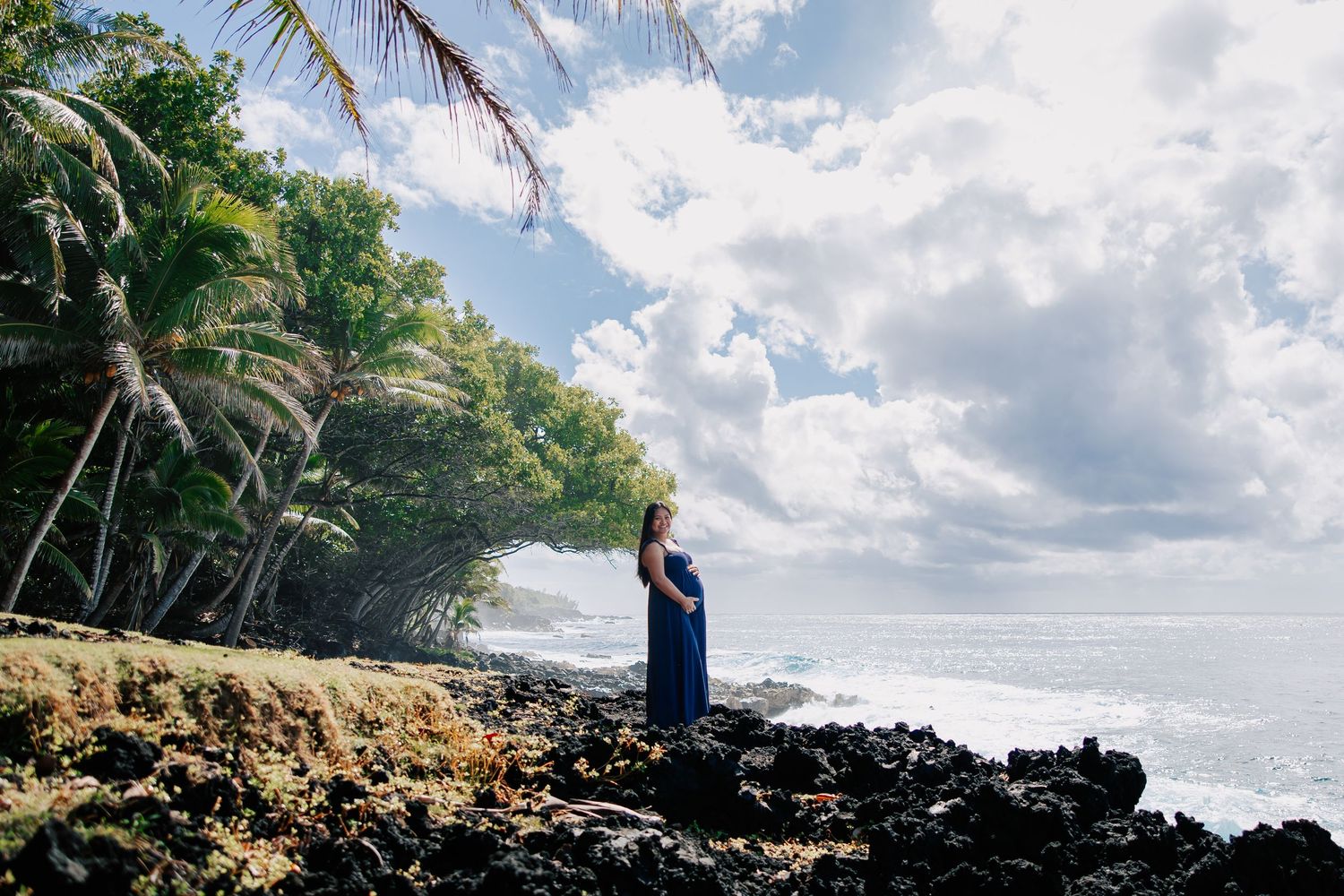 Pregnant woman in blue dress standing on rocky Hawaiian coastline with palm trees and dramatic cloudy sky.