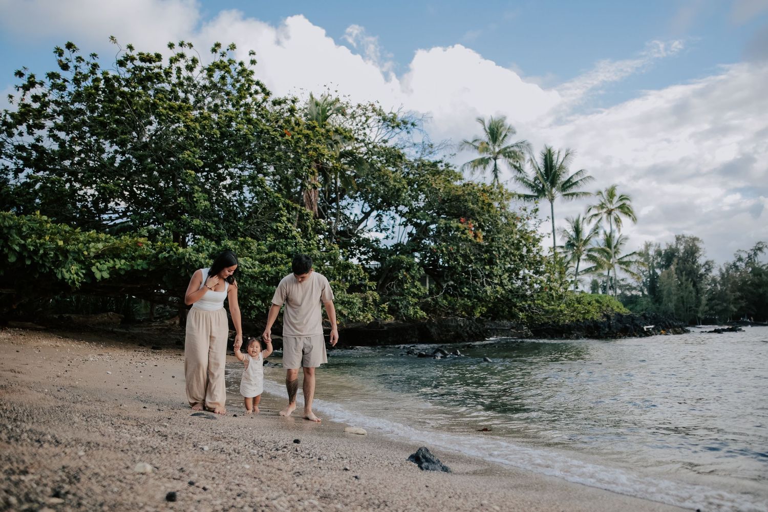 Family of three walking hand-in-hand along a tropical beach with lush palm trees and calm waters.
