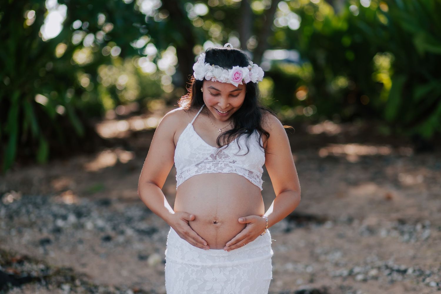 Pregnant woman in white lace dress and floral crown cradling her baby bump outdoors.