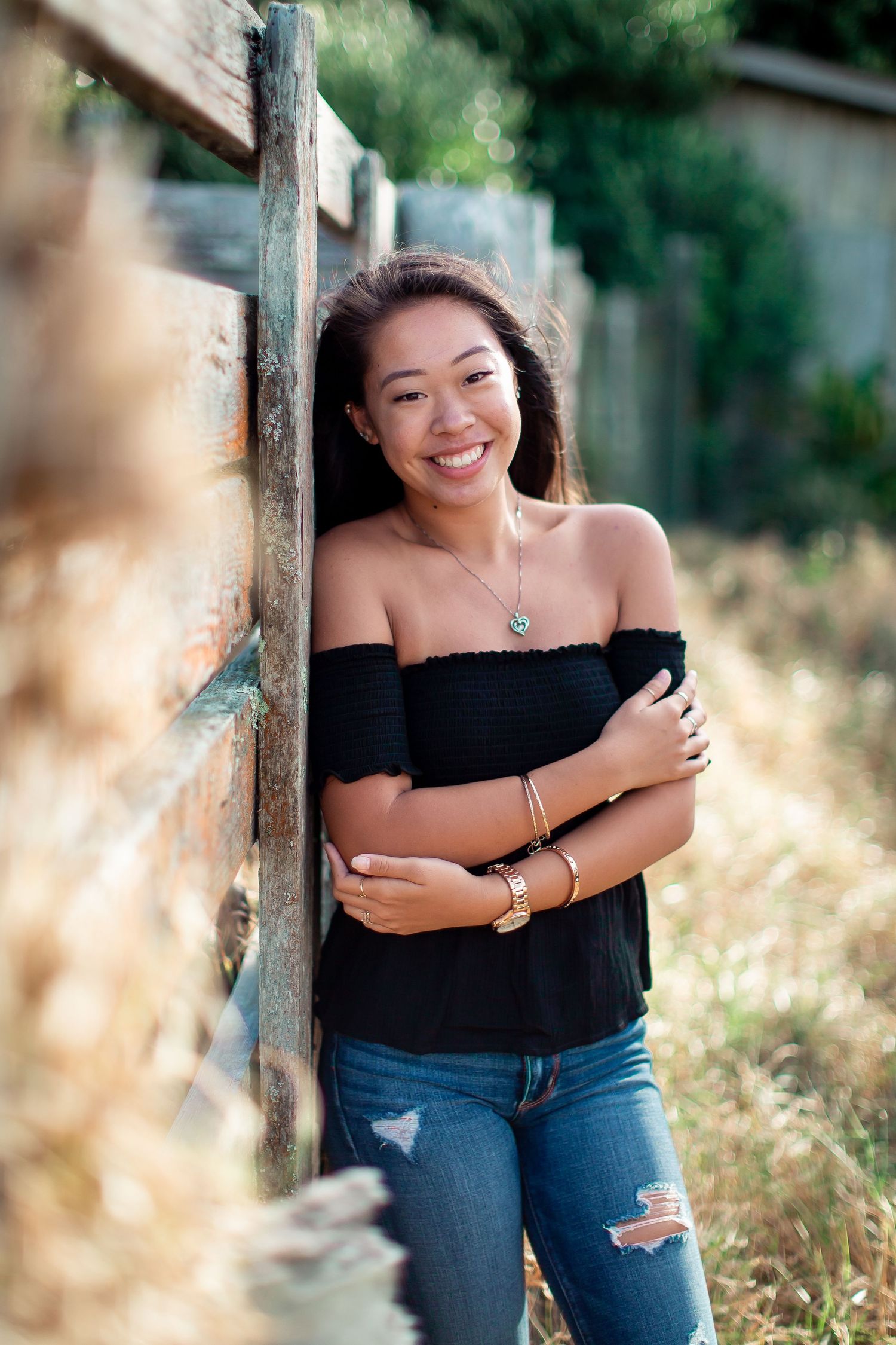 Smiling young woman in black off-shoulder top and ripped jeans leaning against rustic wooden fence outdoors.