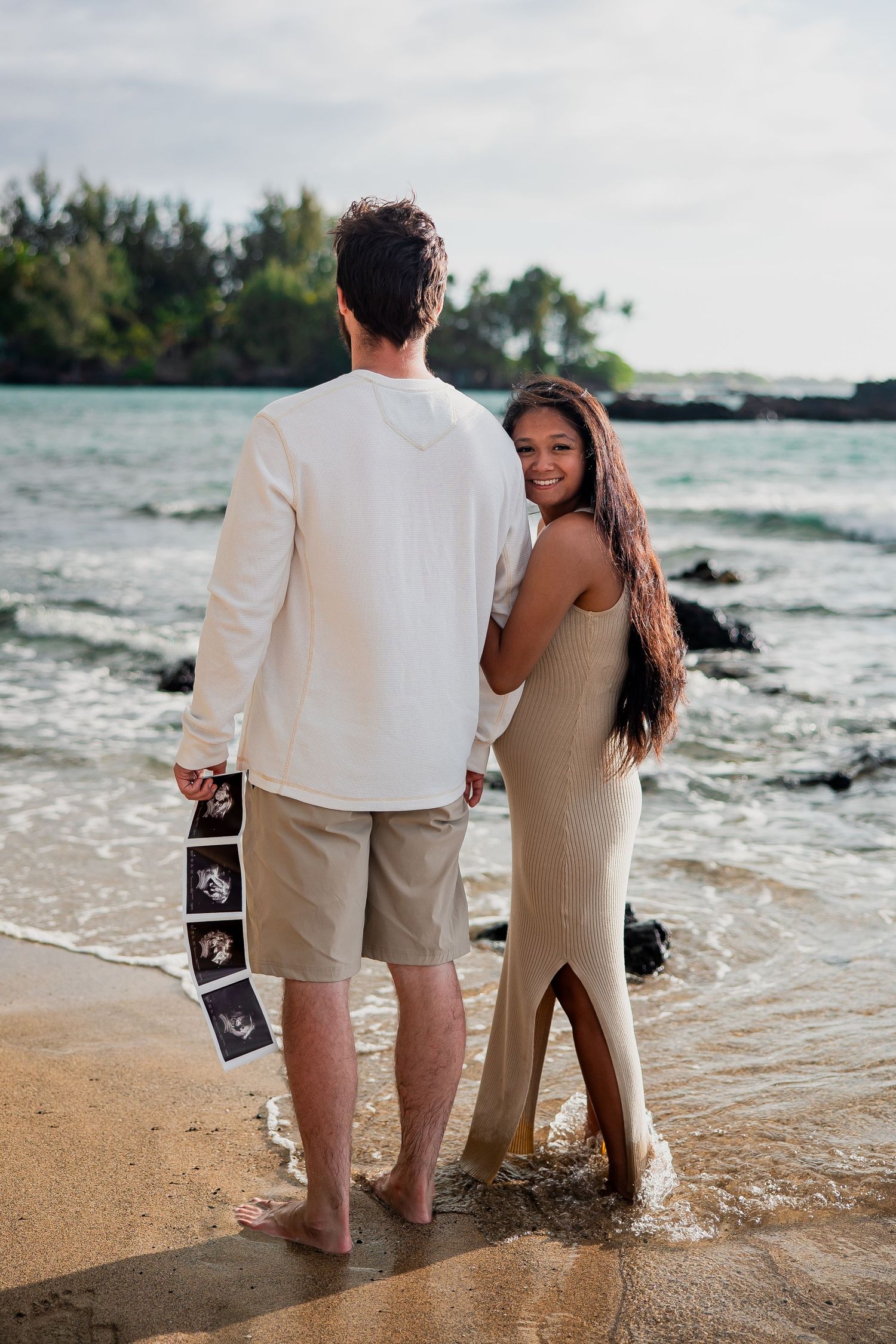 Couple standing on a tropical beach holding ultrasound photos, woman in gold dress smiling, announcing pregnancy.