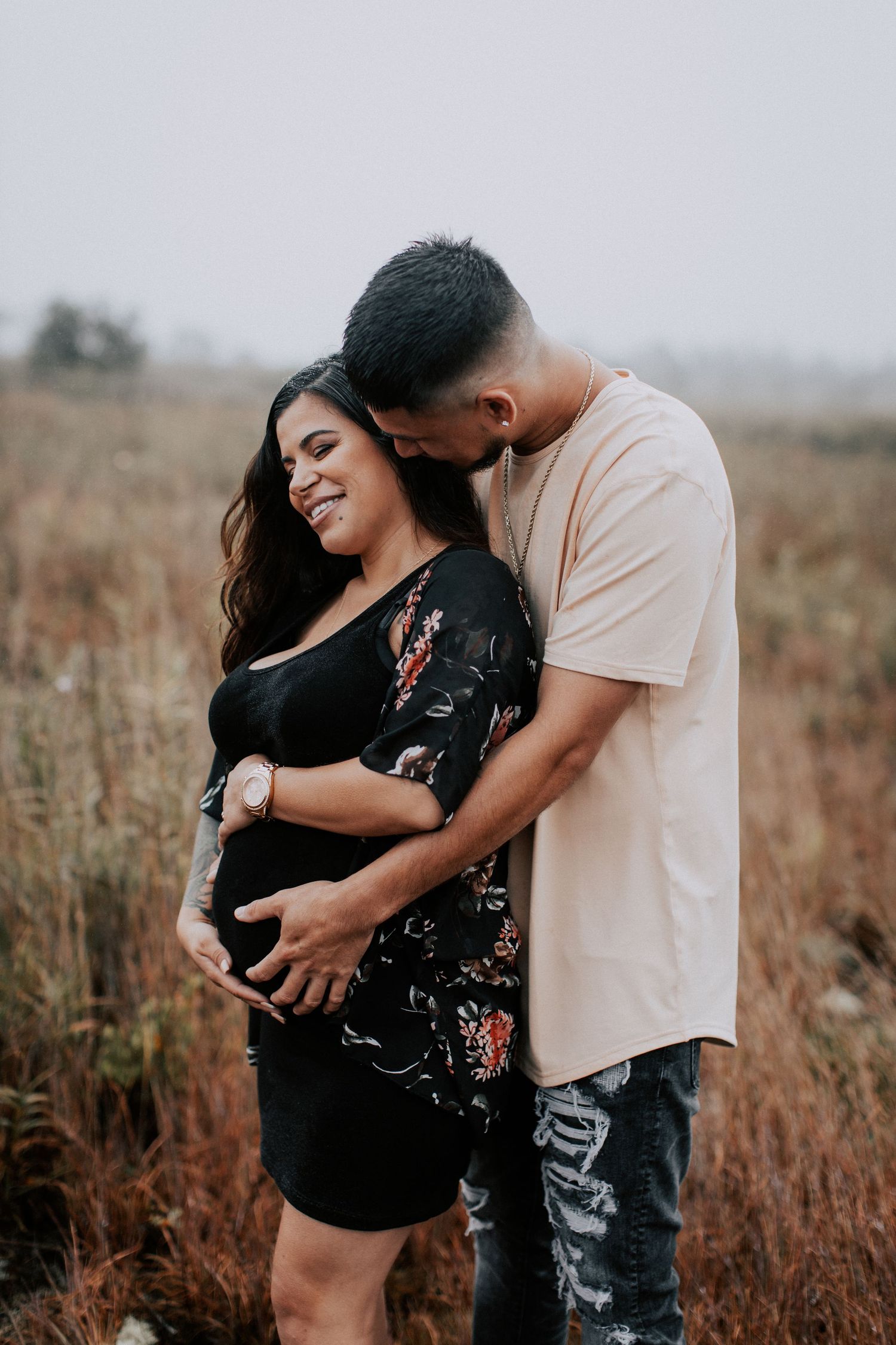 A smiling pregnant woman in a black floral dress stands with her partner in a misty field, both cradling her baby bump.