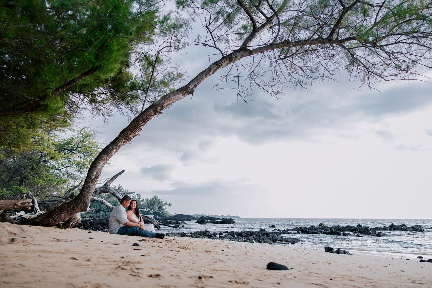Woman sitting under a leaning tree on a rocky Hawaiian beach with dramatic cloudy skies and ocean views.