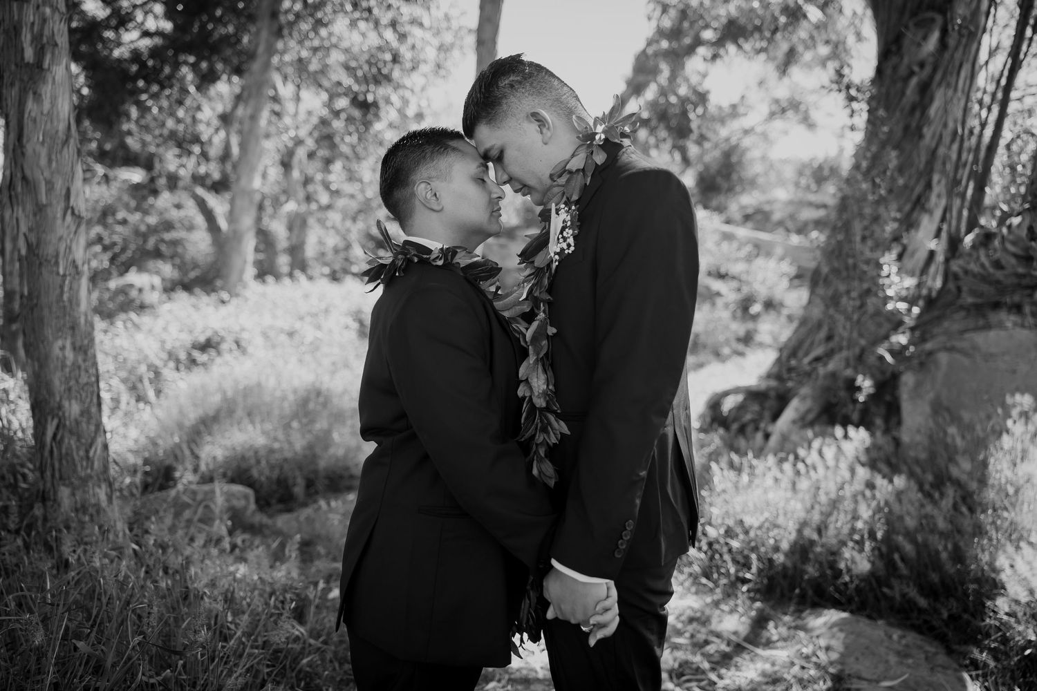 Black and white photo of two grooms in suits sharing an intimate moment outdoors, wearing floral leis.