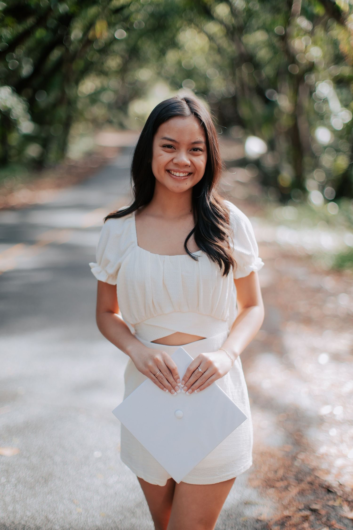 Smiling young woman in white outfit holding graduation cap on tree-lined path.