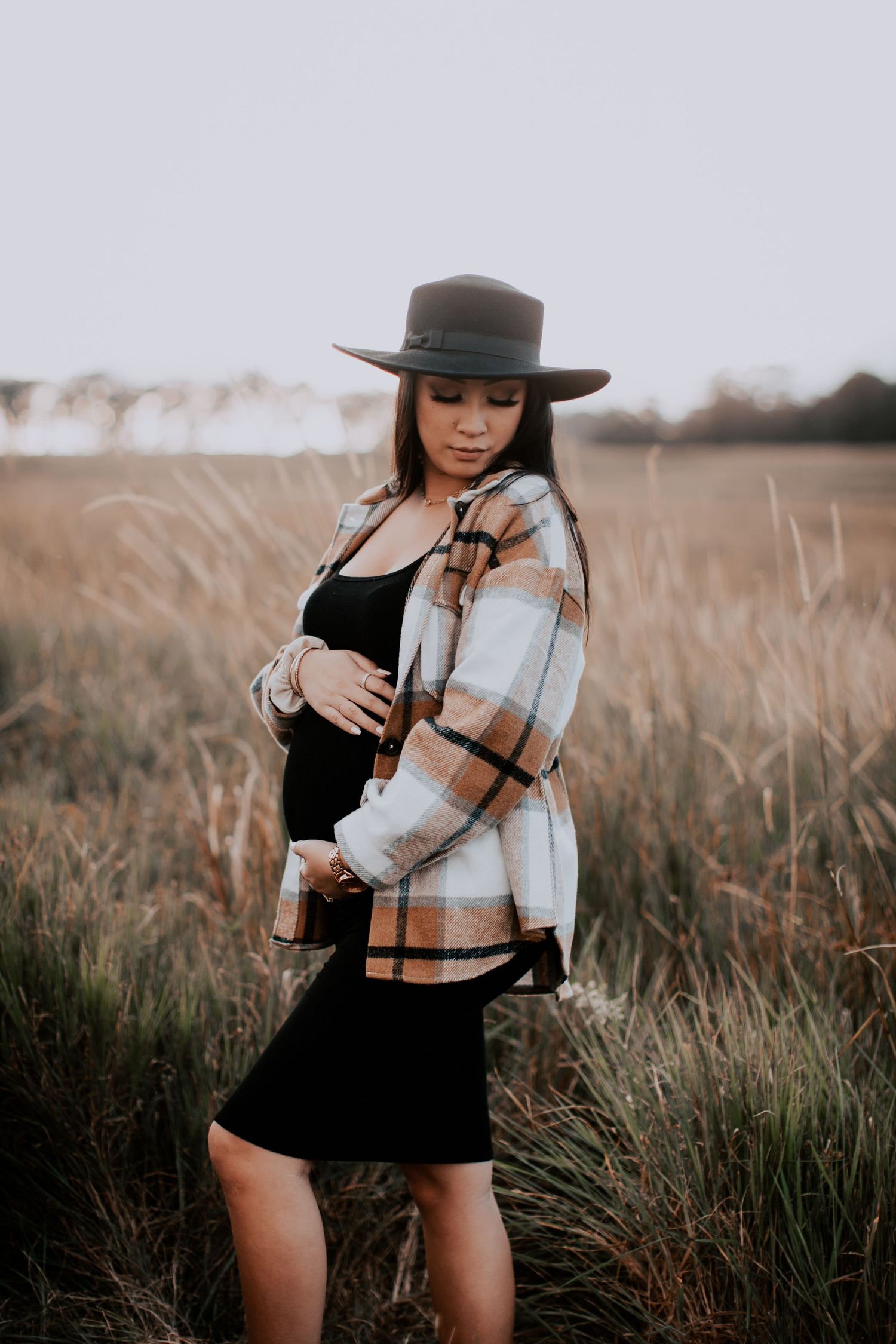 Pregnant woman in plaid shirt, black dress, and wide-brim hat standing in golden field during maternity photo shoot.