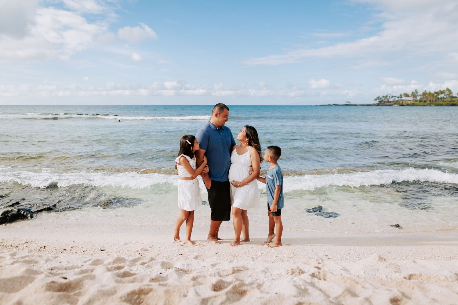 Family of four posing on a white sandy beach in Hawaii with turquoise ocean waves and blue sky behind them.