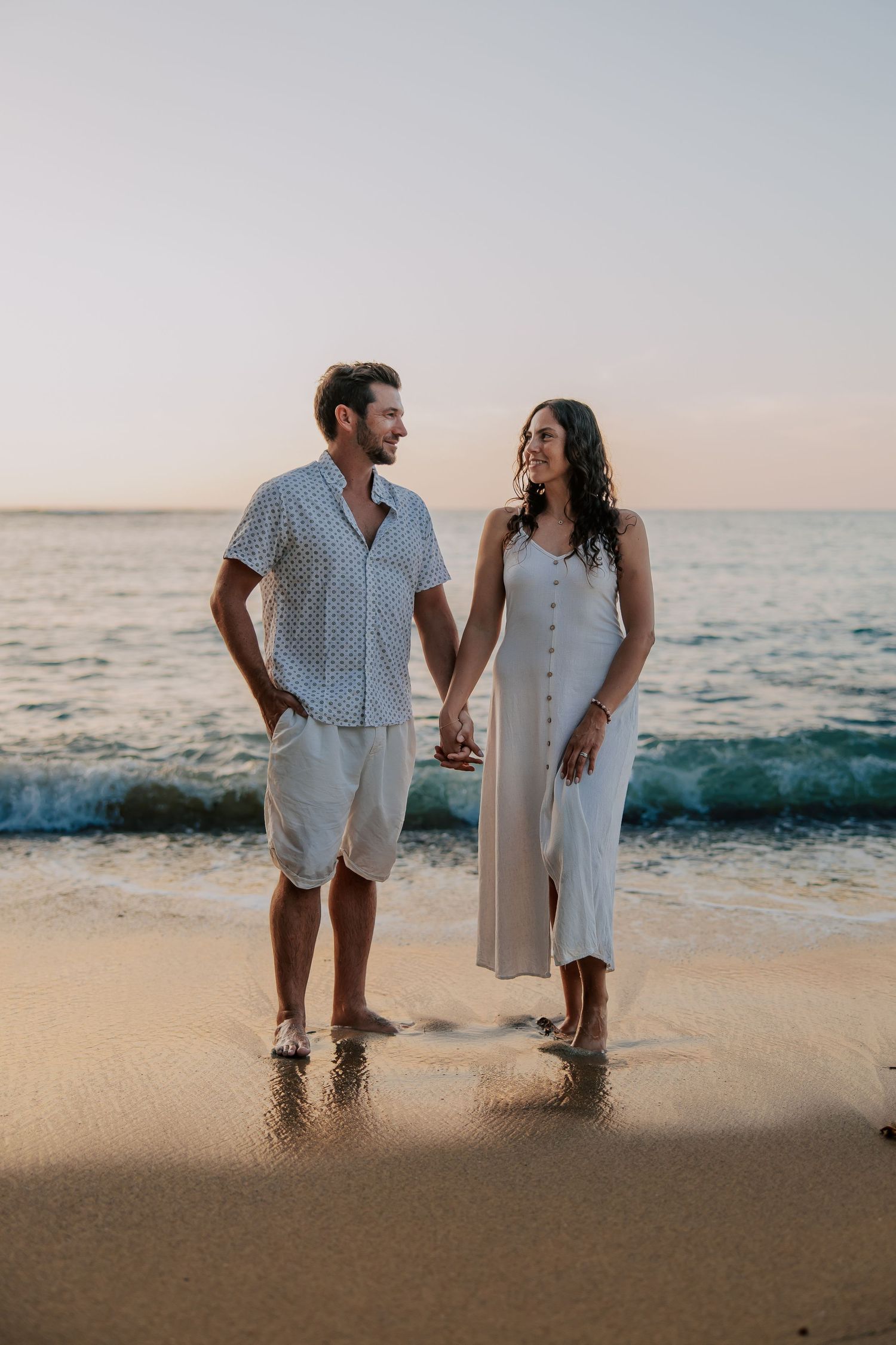 Couple holding hands on a sandy beach at sunset, wearing casual summer outfits, smiling at each other with ocean waves behind.