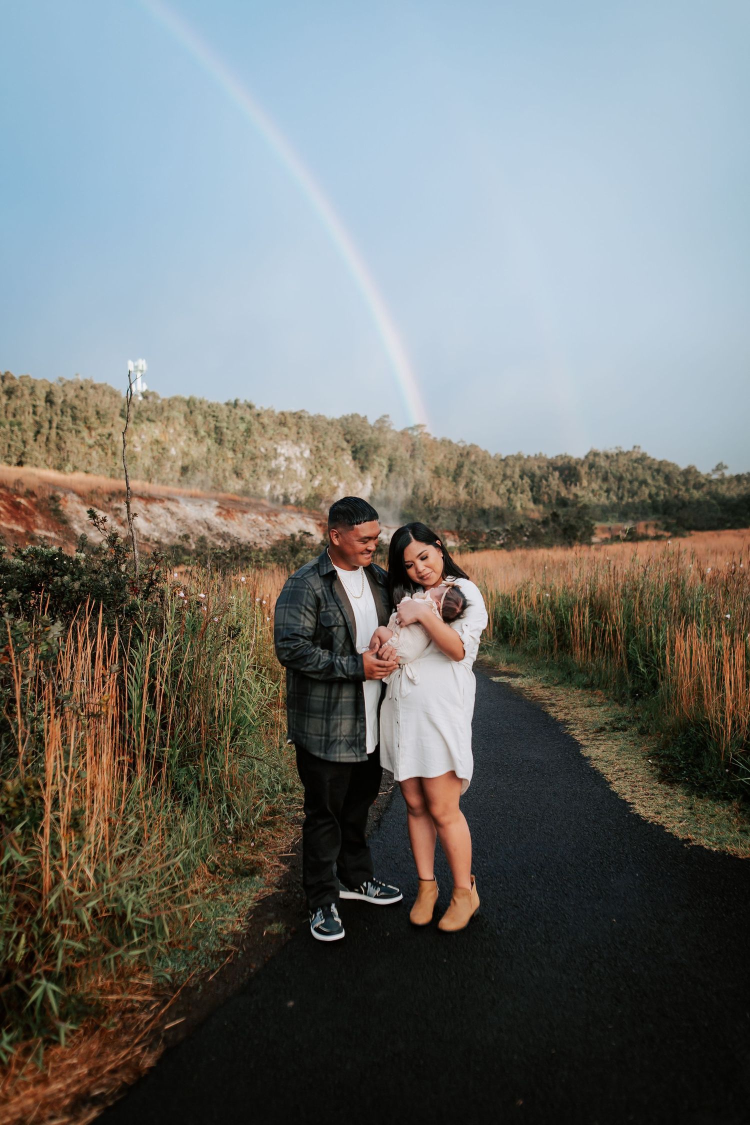 A couple stands on a path holding a newborn baby with a rainbow arching over lush hills behind them.