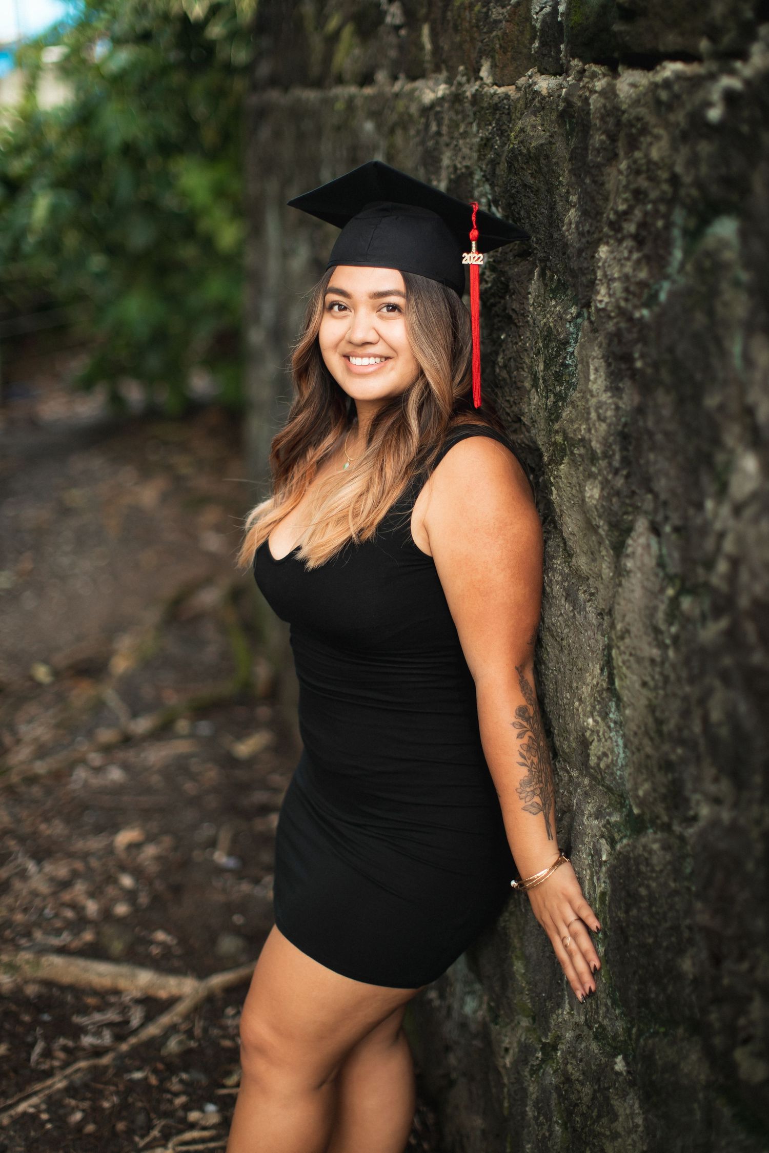 Young woman in black dress and graduation cap smiles against stone wall outdoors.