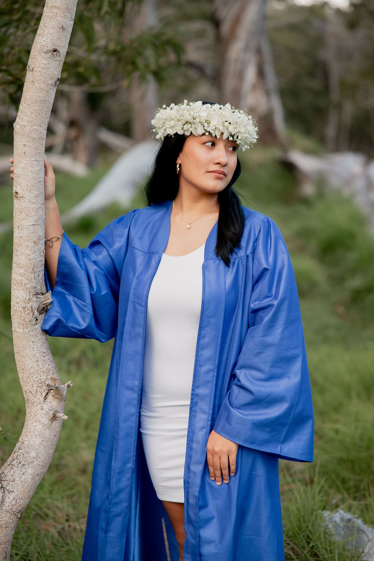 Young woman in blue graduation gown and white floral crown poses outdoors among trees.