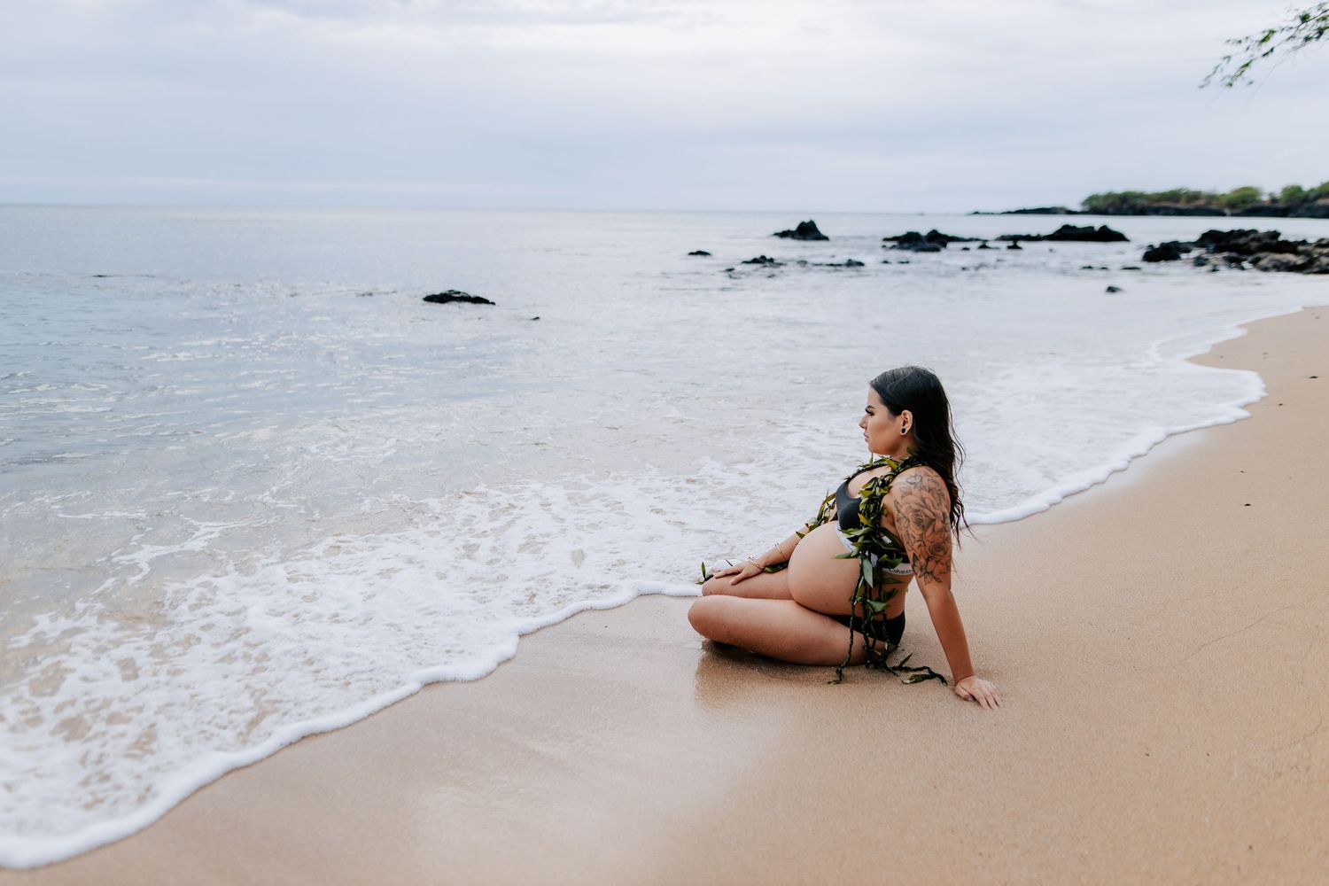 A woman in a floral bikini sits peacefully on a sandy beach, gazing at calm ocean waves under an overcast sky.