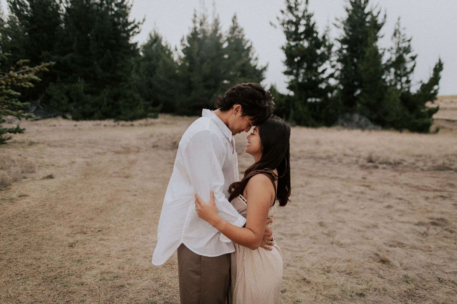Couple sharing a romantic kiss outdoors in a field with evergreen trees in the background.