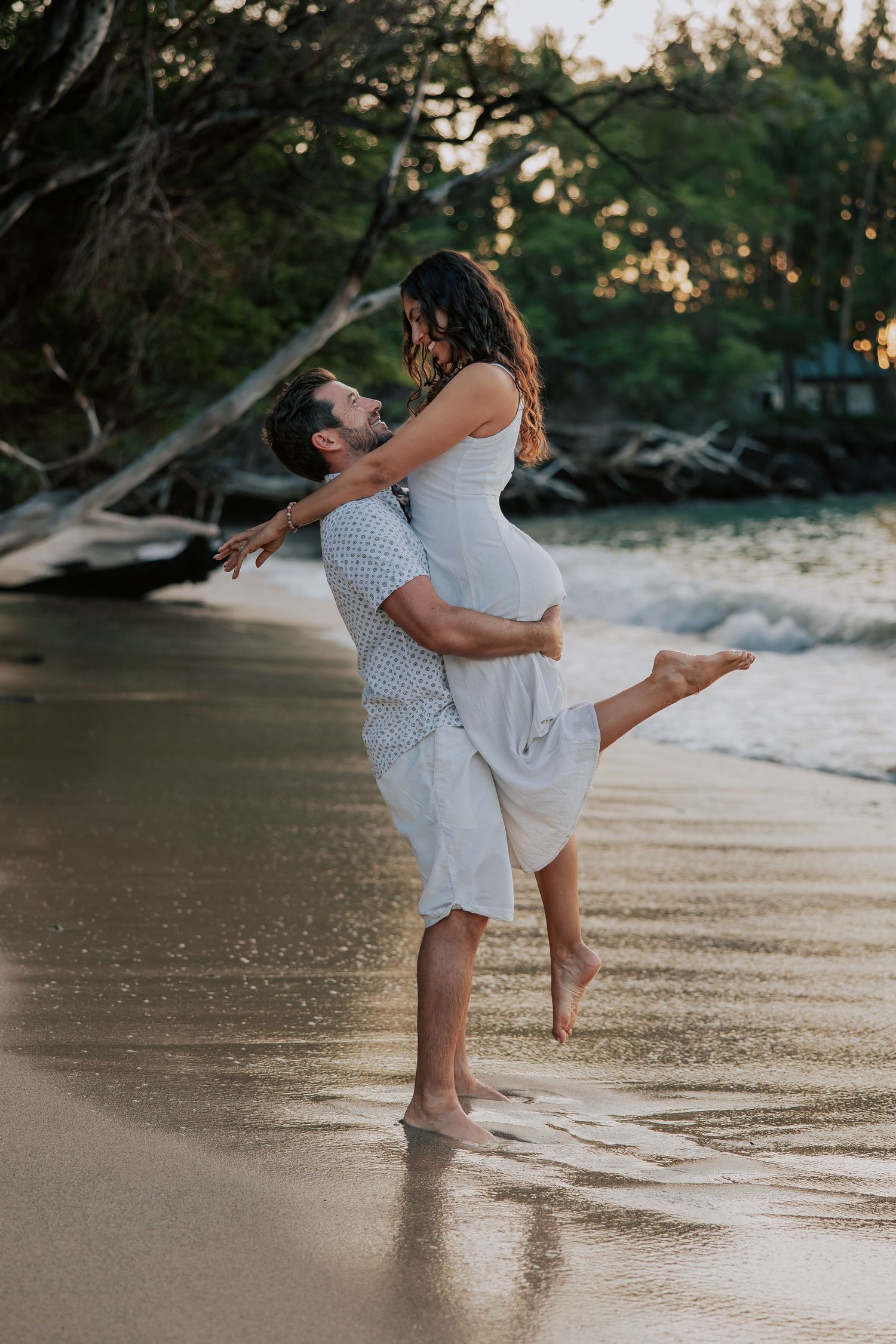 A couple shares a romantic beach moment as a man lifts a woman in his arms at sunset near the shoreline.