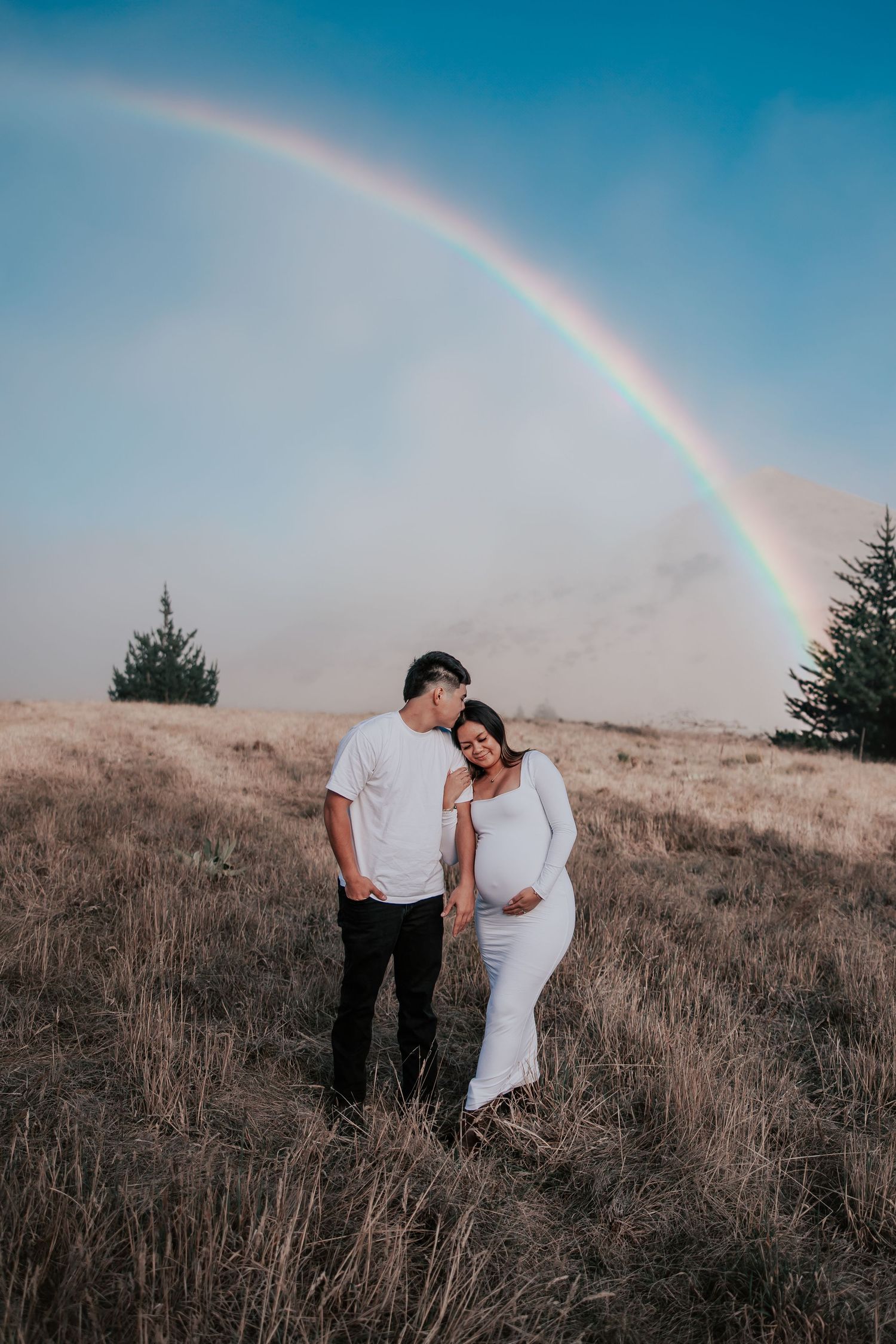 Couple in white outfits sharing a tender moment in a golden field under a vibrant rainbow during a maternity photoshoot.
