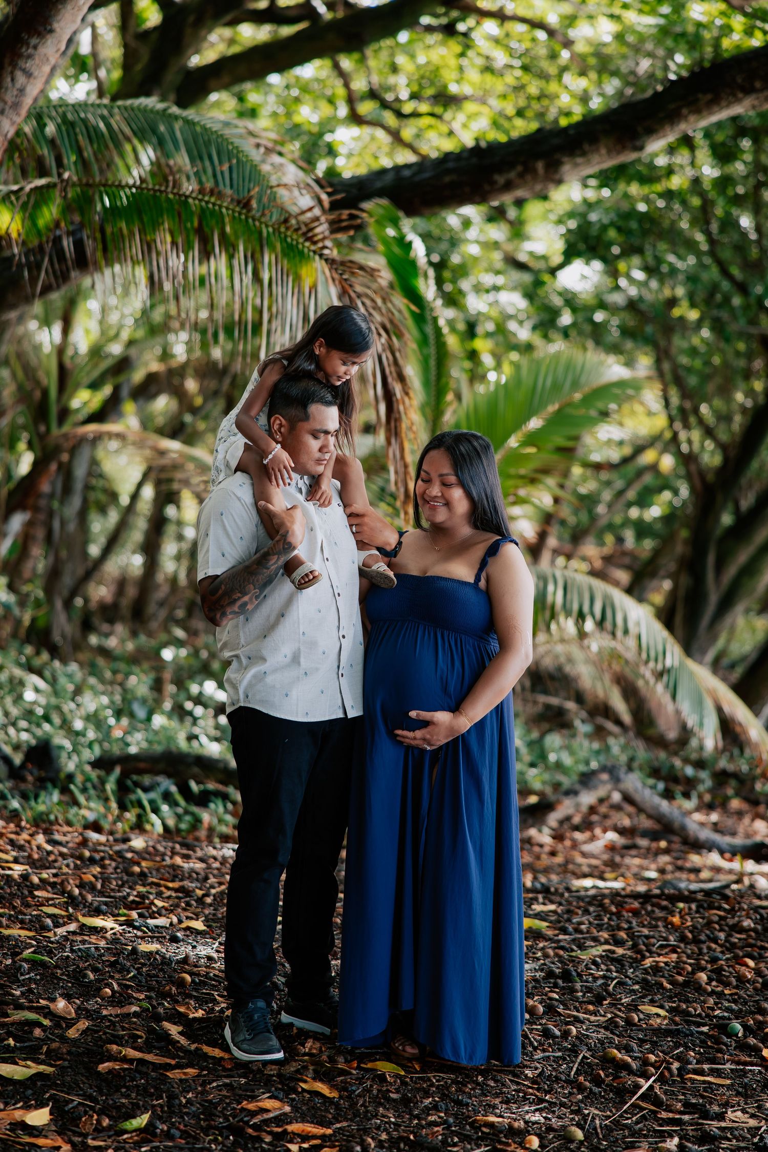 Pregnant woman in navy blue dress with family in tropical garden setting for maternity photo shoot.