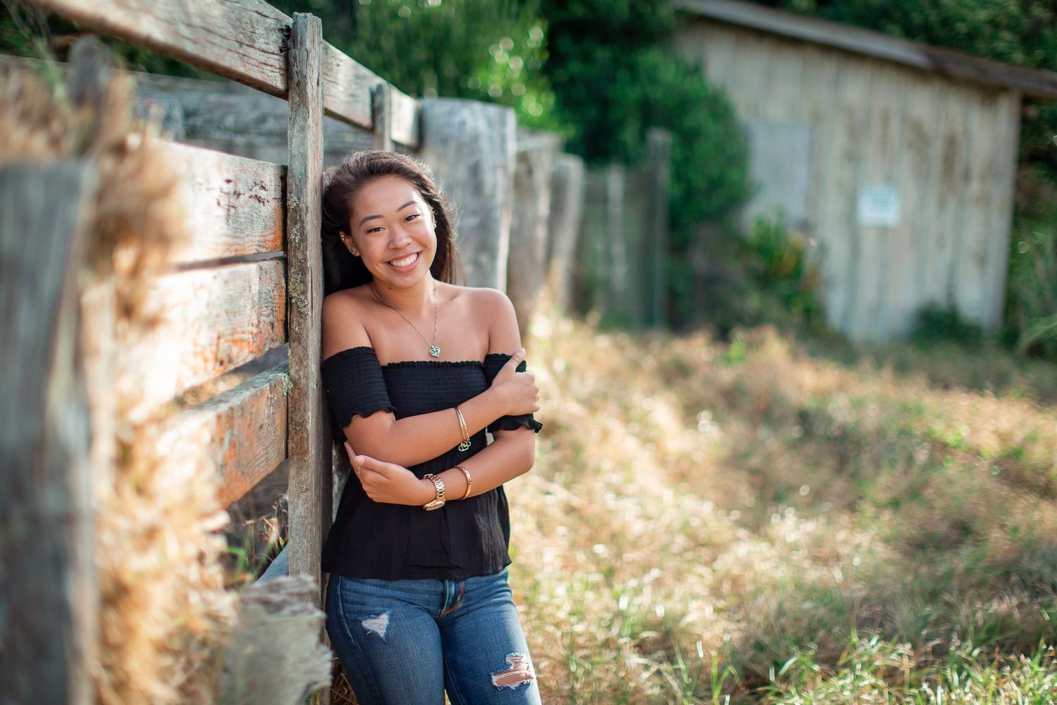 Young woman in black off-shoulder top and ripped jeans leaning against a wooden fence outdoors.