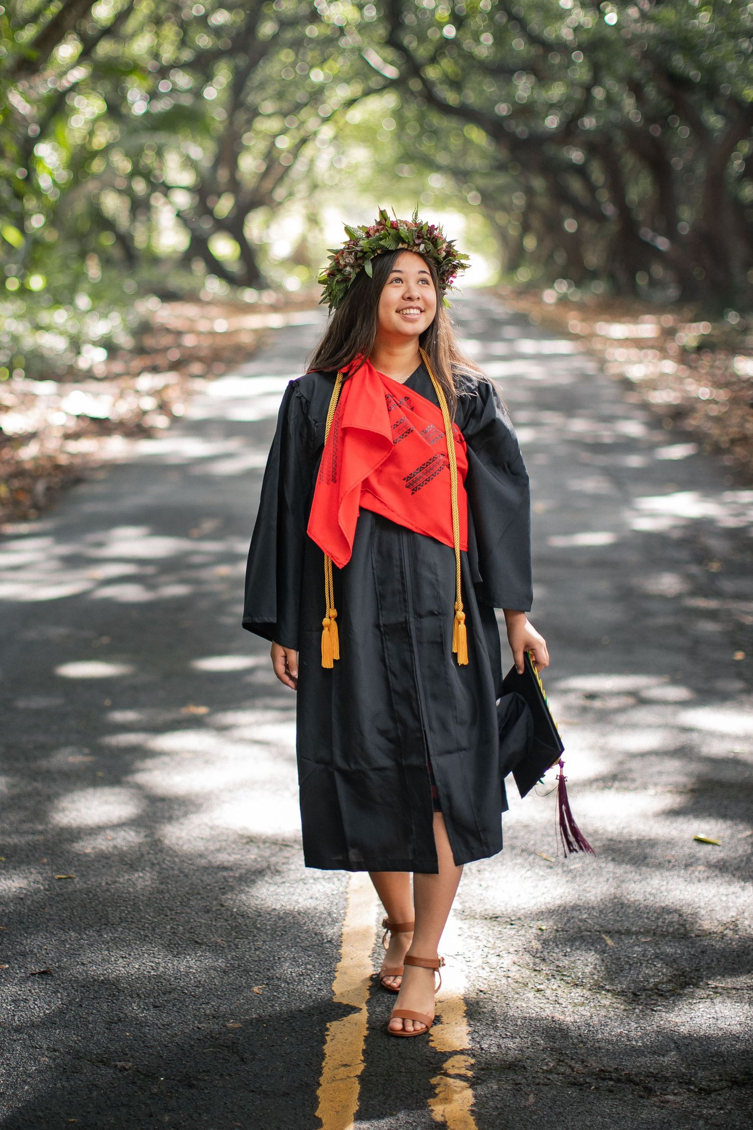 Smiling graduate in black cap and gown with red stole and floral crown walks on a tree-lined road.