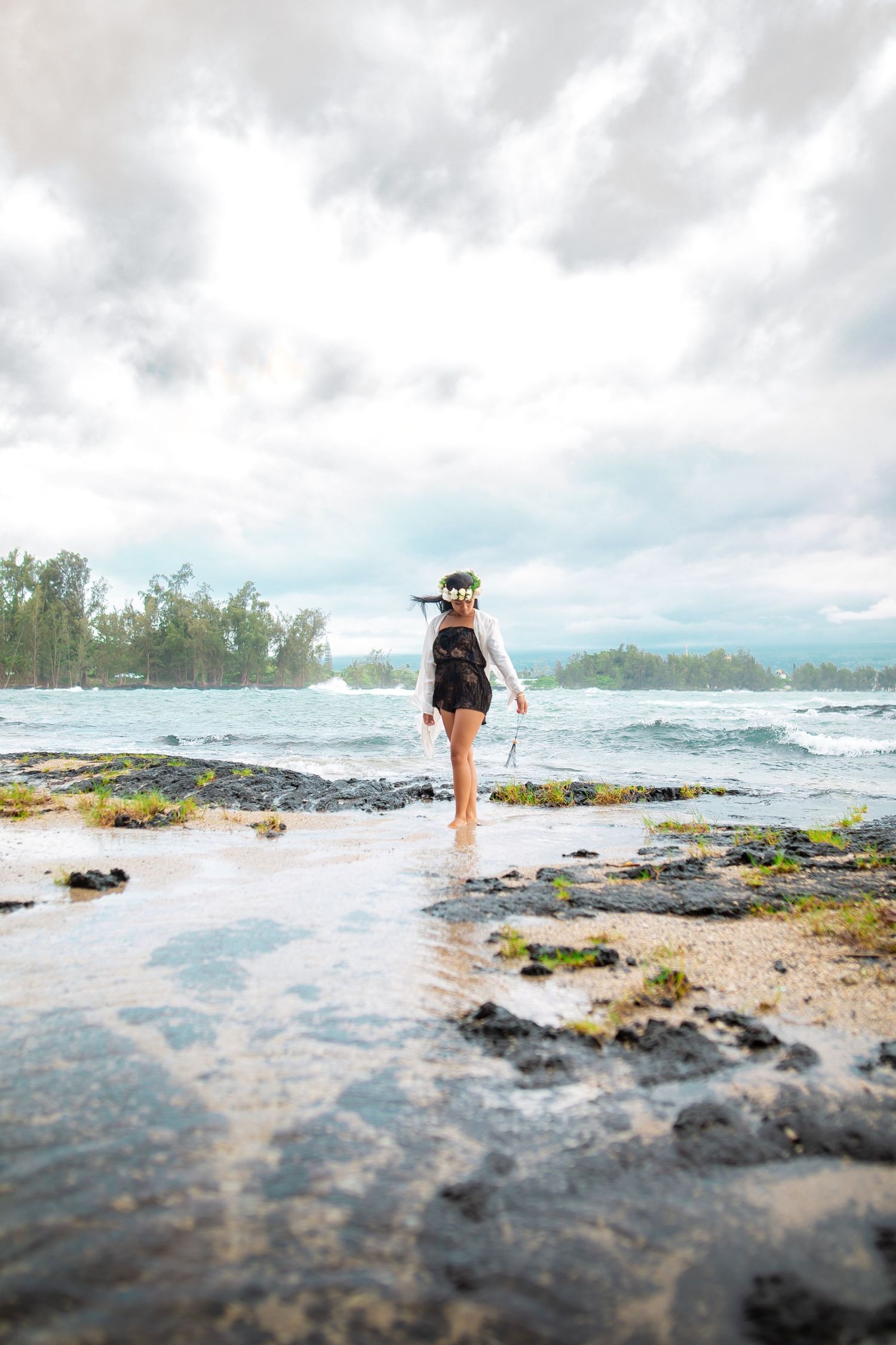 Woman in black swimsuit and floral crown walks along rocky Hawaiian shoreline with dramatic cloudy skies.