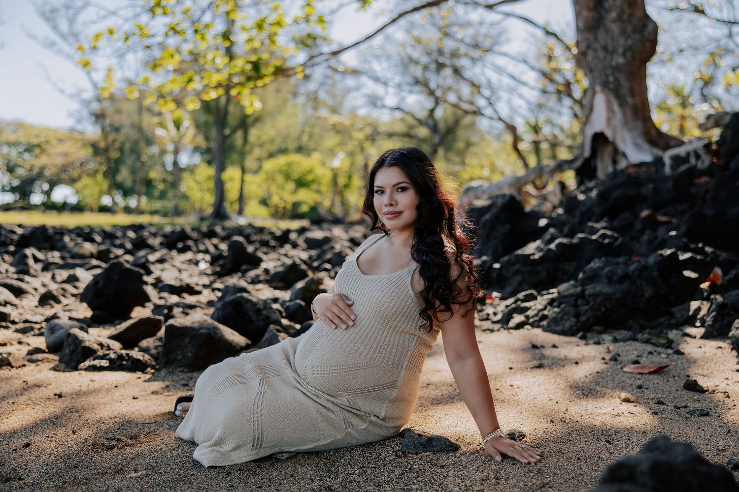 Pregnant woman in a white maxi dress sitting on sandy beach with lava rocks and trees in a maternity photo shoot.