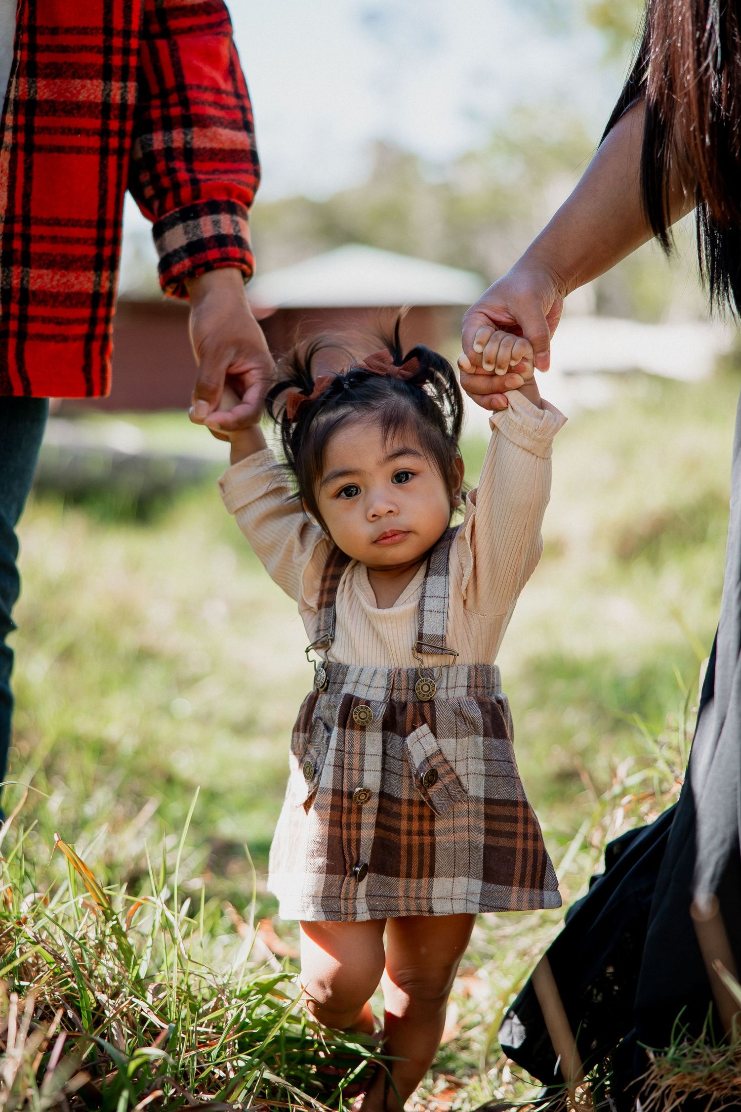 Toddler girl in plaid overalls held by two adults while walking outdoors on green grass.