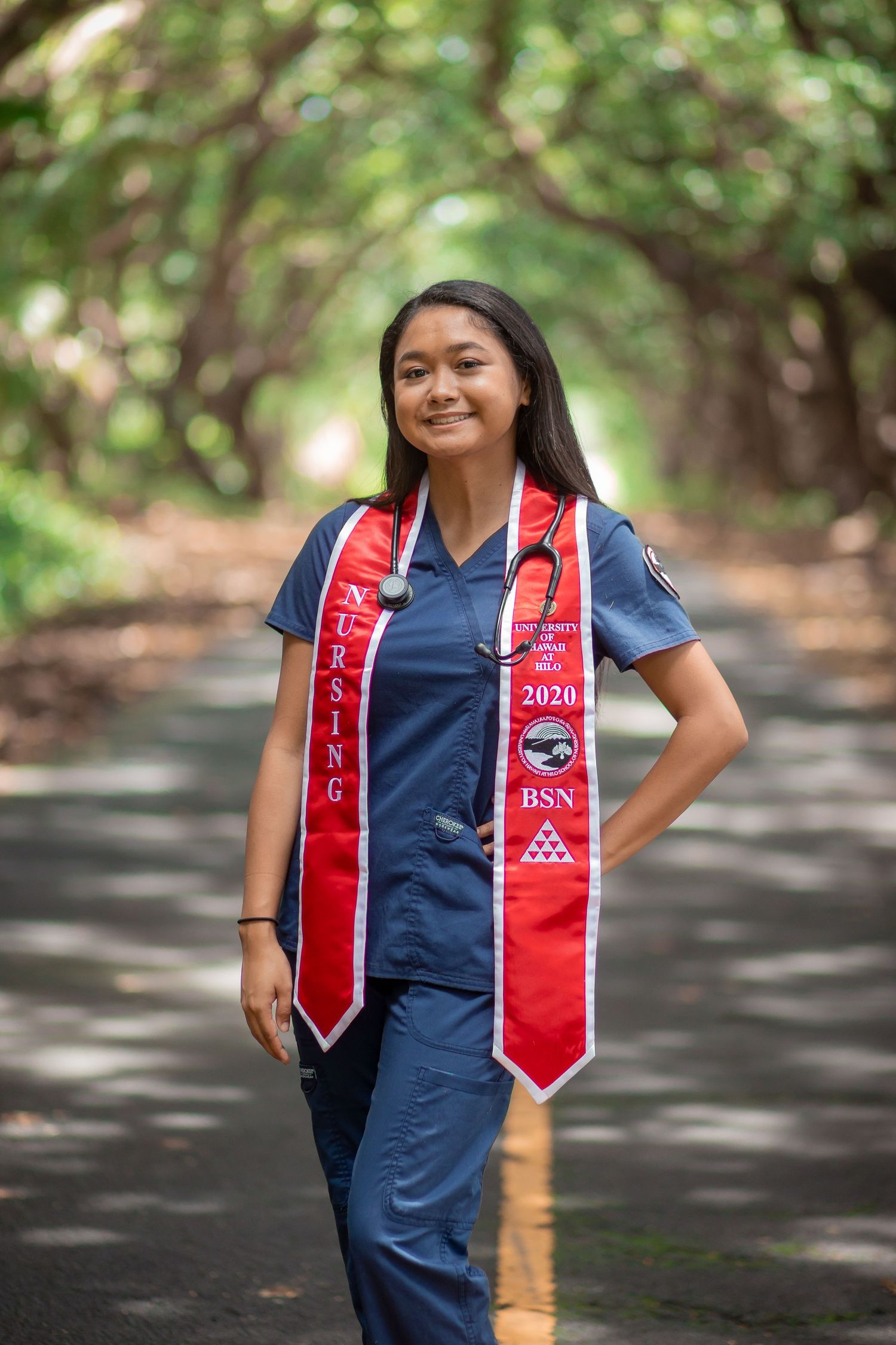 Nursing graduate in blue scrubs with red BSN 2020 graduation stoles and stethoscope poses on tree-lined road.