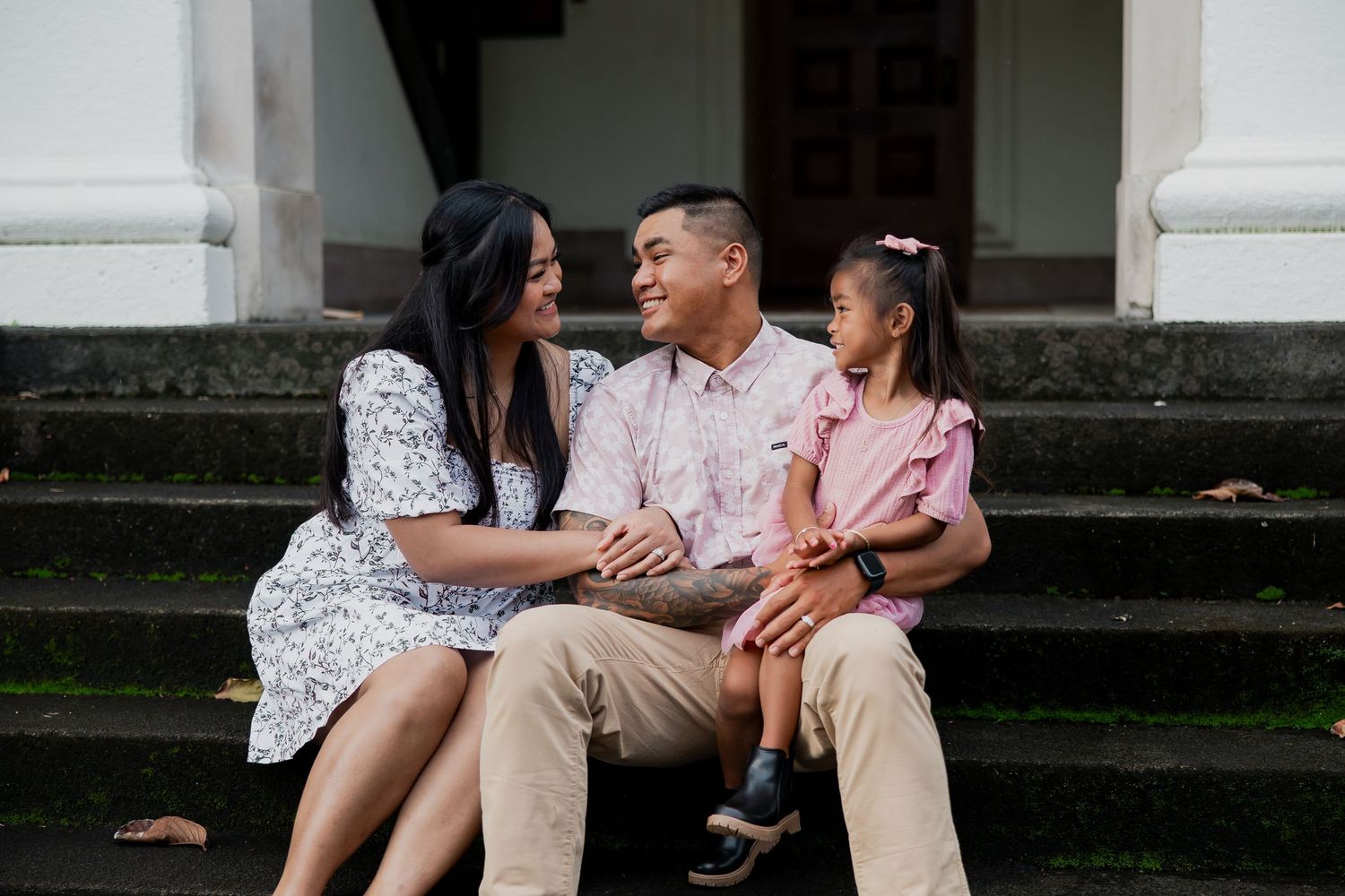 Happy Asian family of three sitting on steps, parents smiling at cute daughter in pink dress.