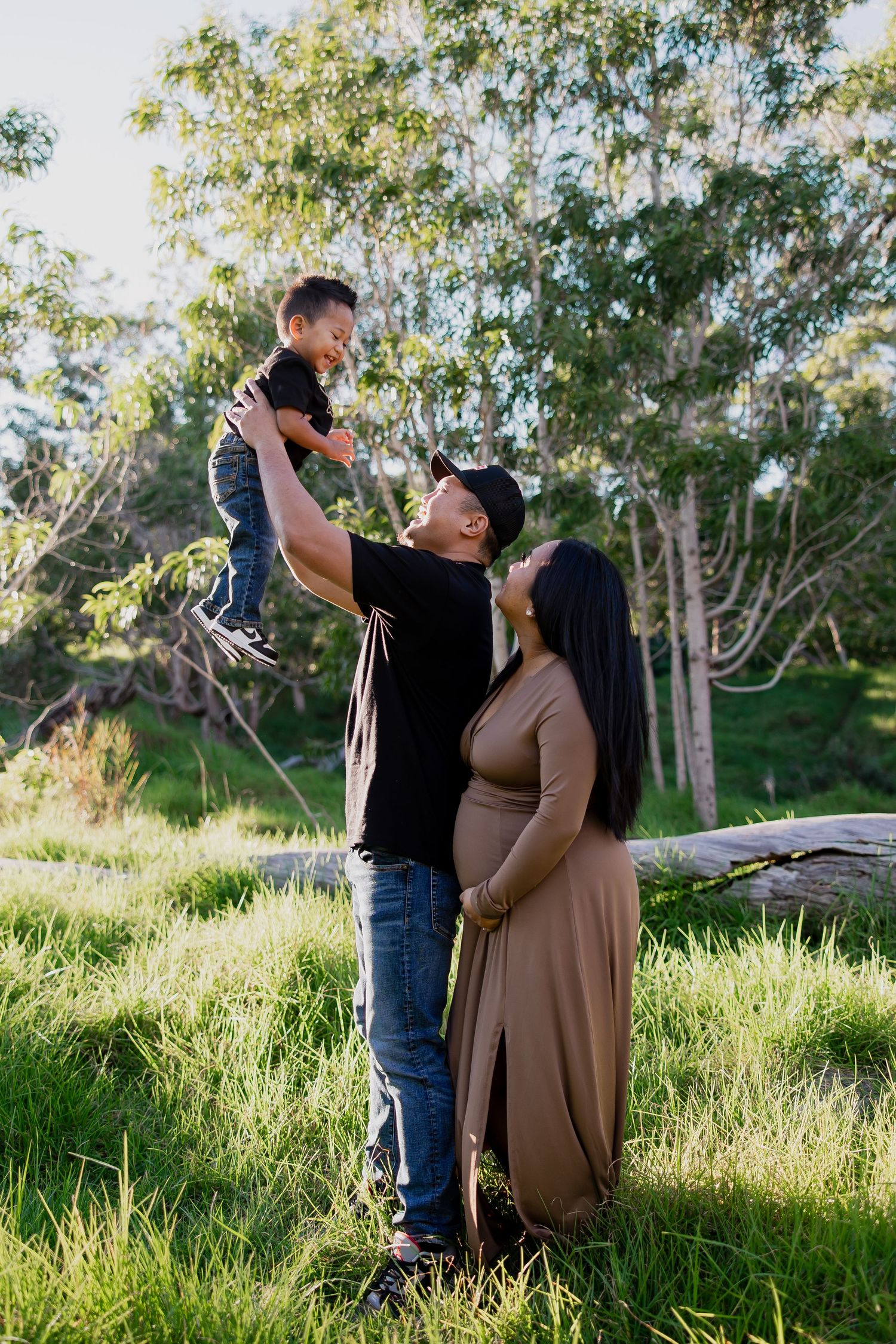 A man lifts a young child while a pregnant woman in a tan dress stands beside them outdoors.