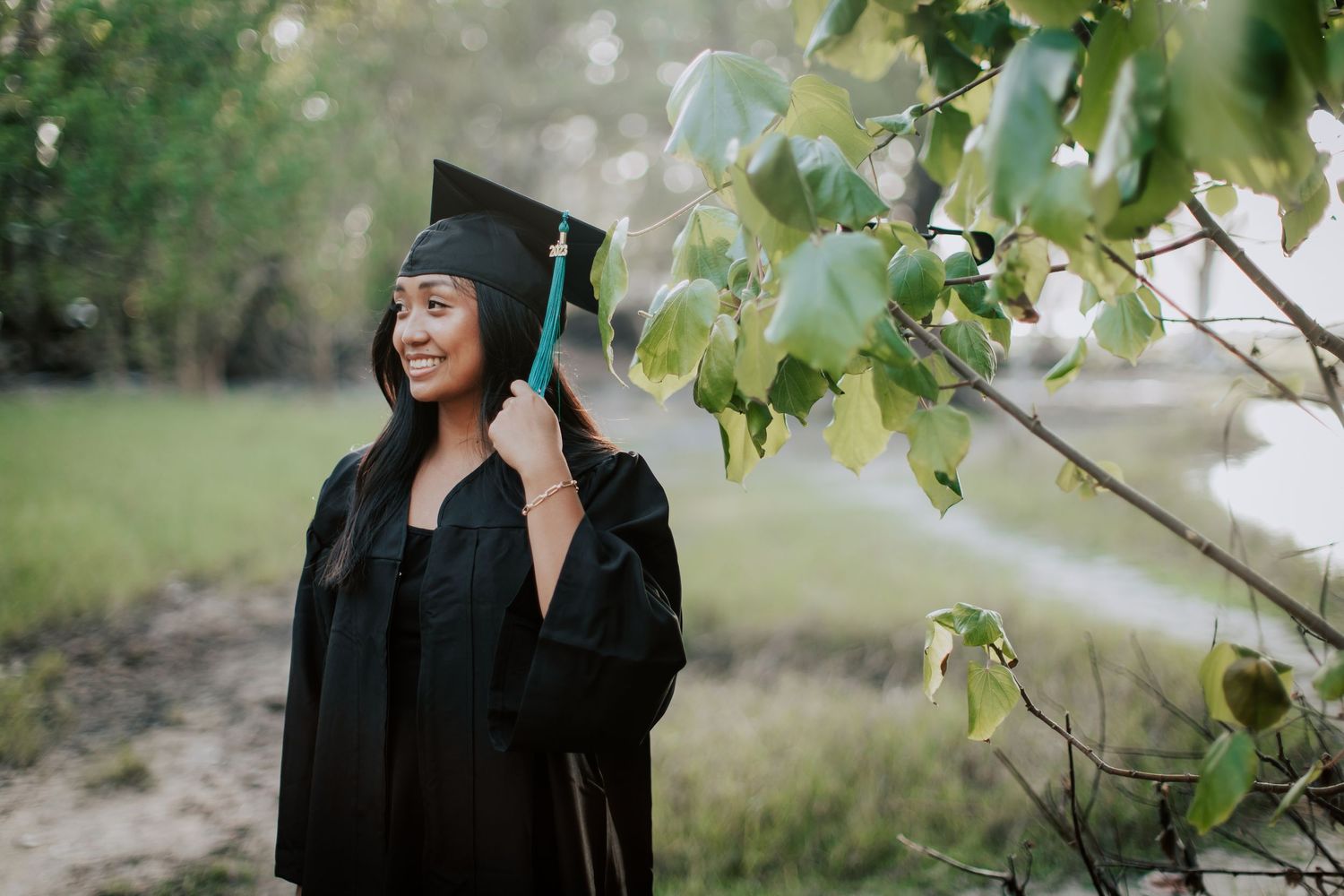 Smiling graduate in black cap and gown posing outdoors among green trees and nature backdrop.