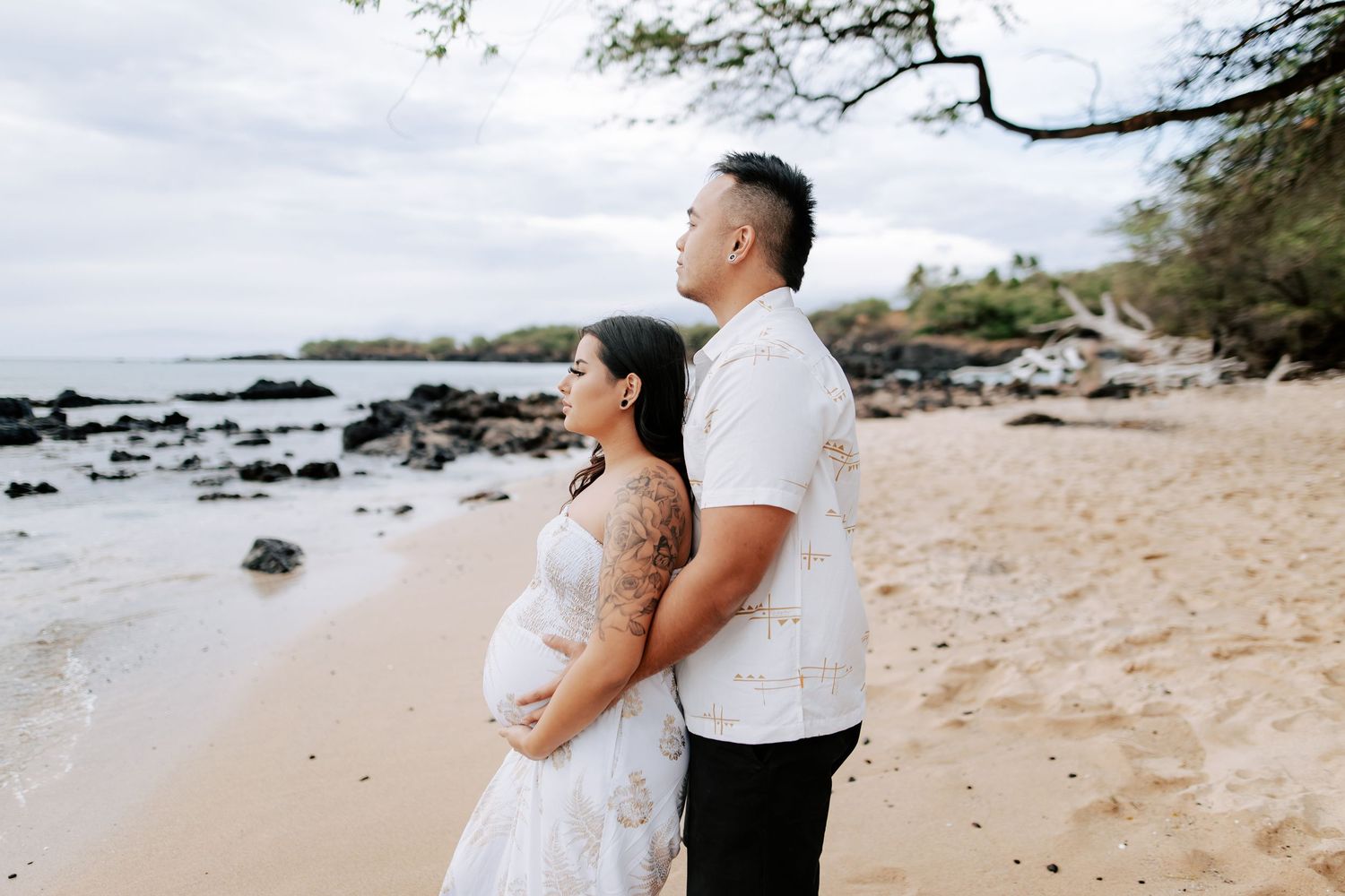 Pregnant couple standing back-to-back on a sandy Hawaiian beach, wearing white outfits, with rocky shoreline in background.
