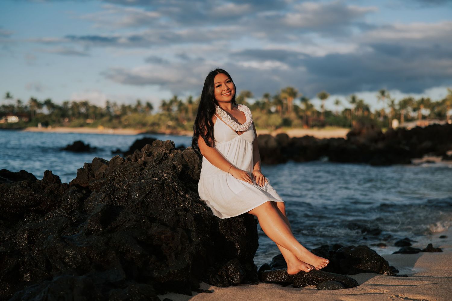 Woman in white dress sitting on lava rocks at a Hawaiian beach during golden hour sunset.