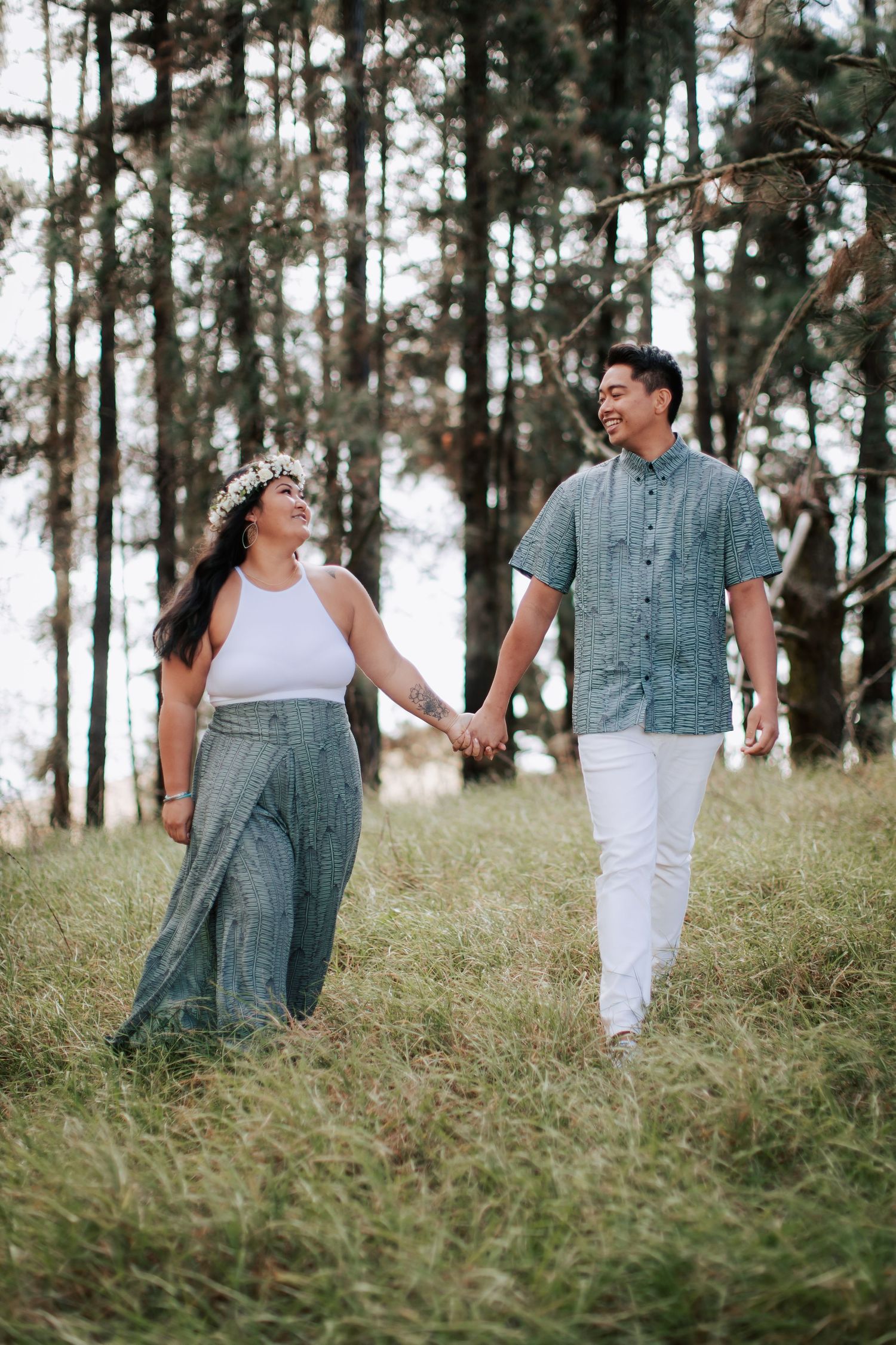 A couple holding hands and walking through a grassy forest, woman wearing floral crown and wide-leg pants, man in blue shirt.