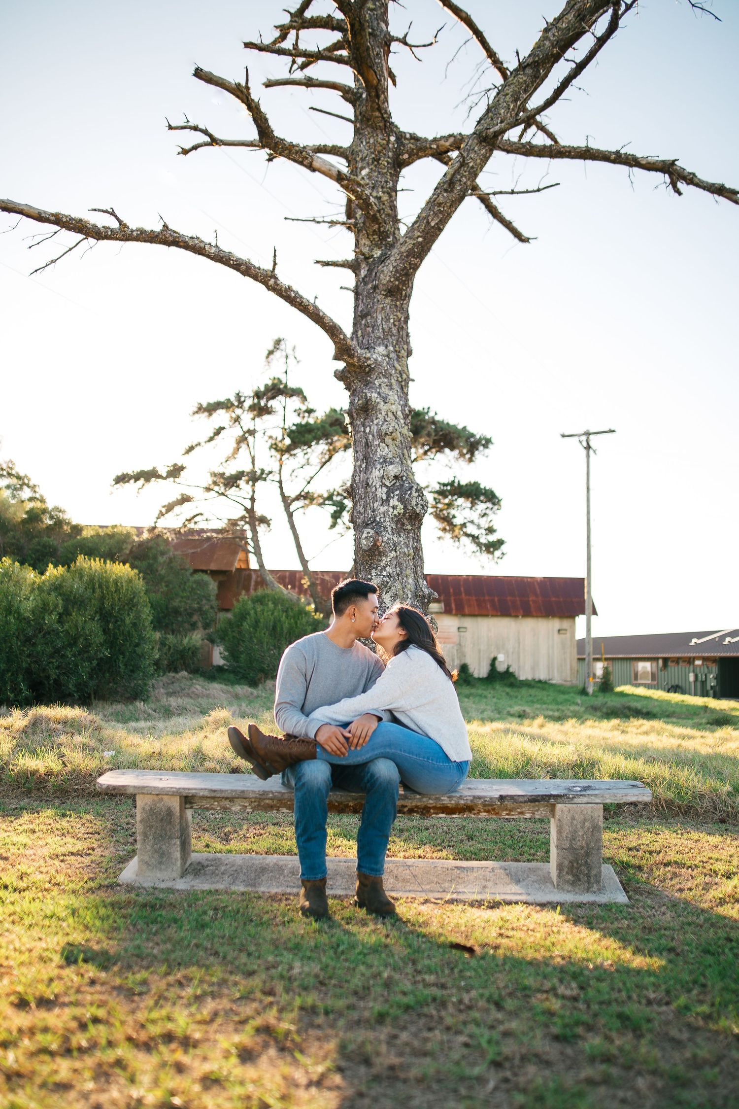 Couple sharing a romantic moment on a stone bench under a bare tree in a rustic outdoor setting.