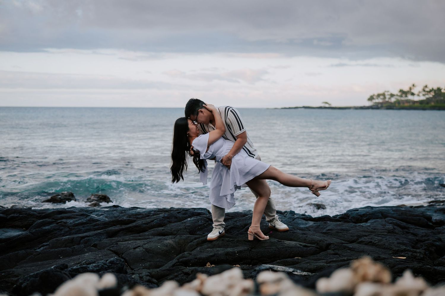 Couple sharing a romantic kiss on black lava rocks by the ocean in Hawaii, with turquoise waves and green palms.